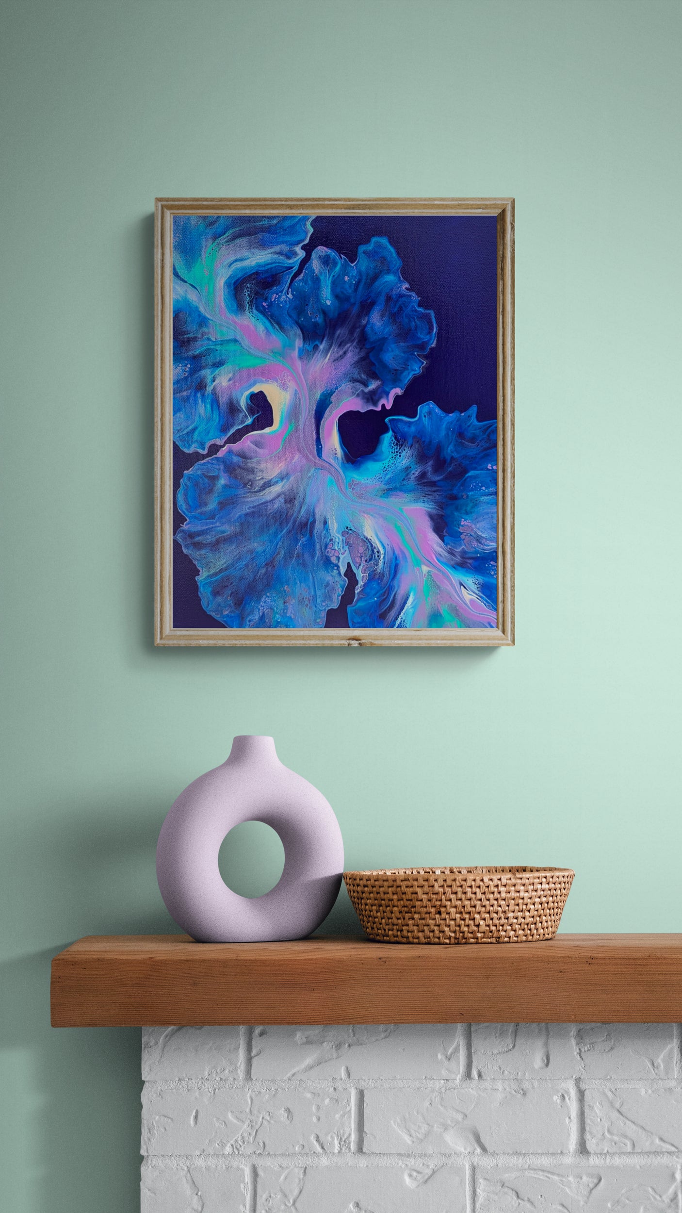 Framed abstract fluid painting titled Bioluminescence in a cozy, modern room. The artwork features vivid neon blues, purples, and greens flowing across a dark navy background, hung on a mint green wall above a wood mantel with a lilac sculptural vase and woven basket.