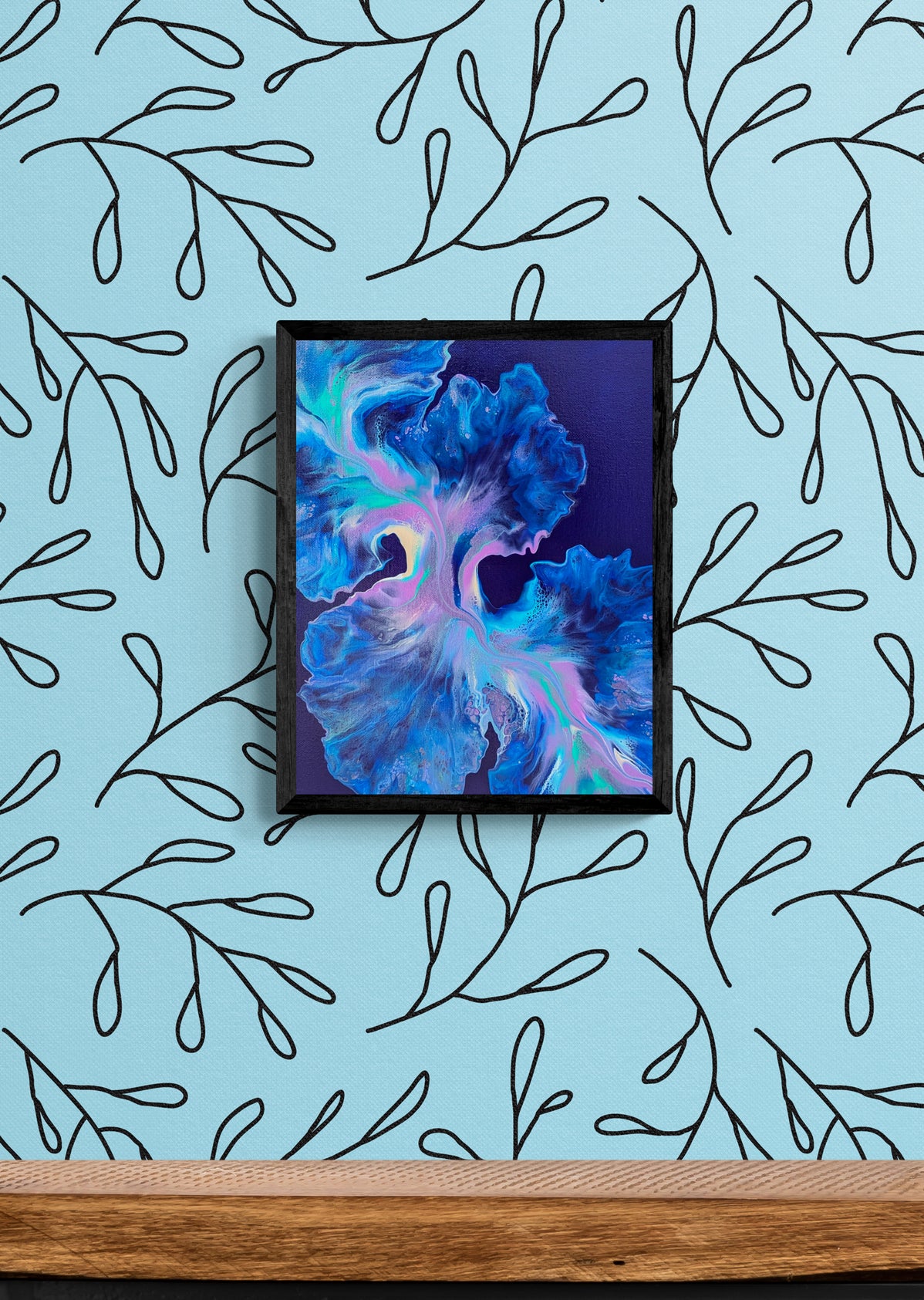 Framed abstract painting titled Bioluminescence in a vertical layout. The painting features glowing neons and deep blues on a navy background, set against a light blue wallpaper with a black hand-drawn vine pattern above a natural wood shelf.