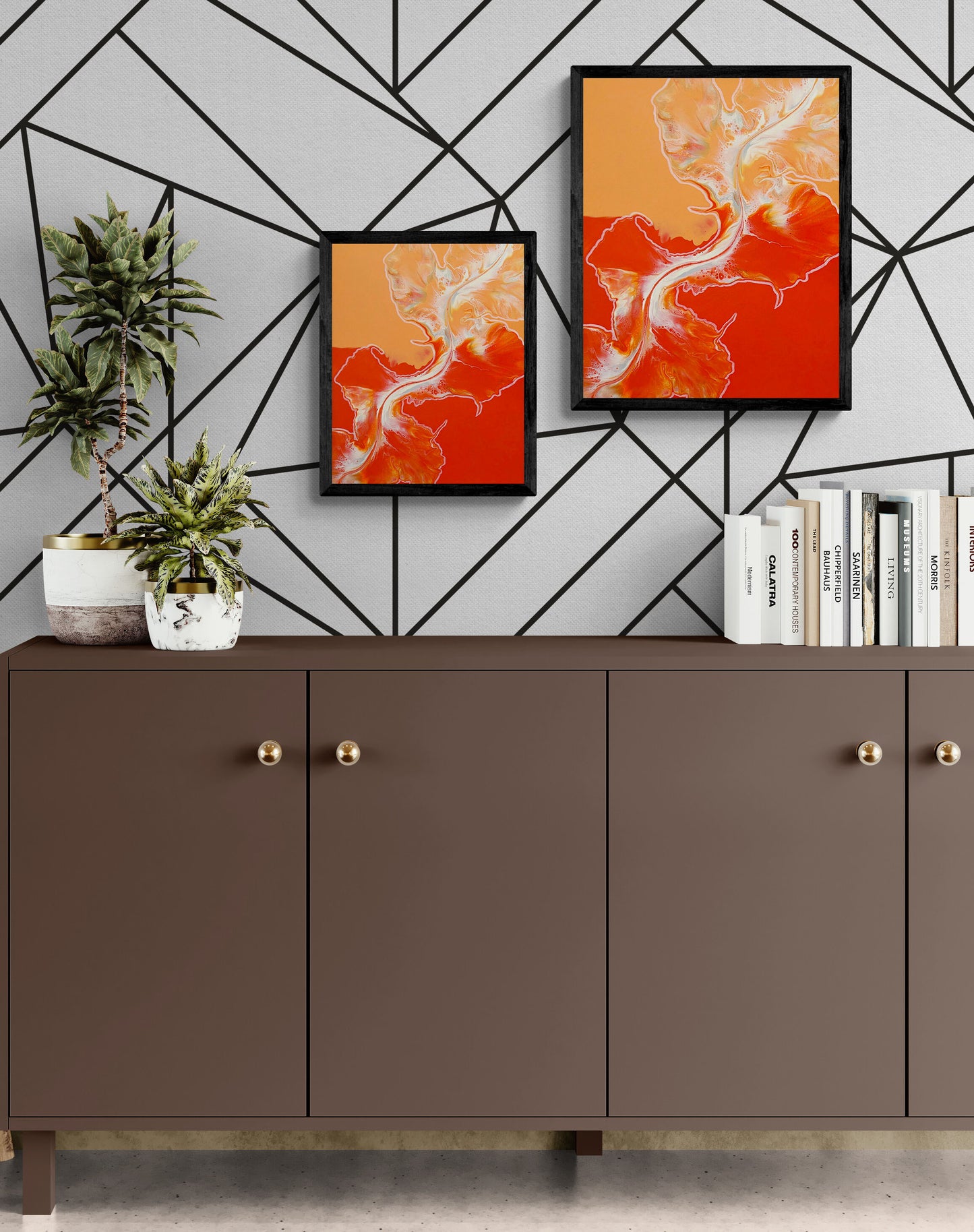 Two framed acrylic fluid art prints of Cross the Line, one small and one large, hung side by side on a geometric patterned wall above a modern brown credenza with plants and books.