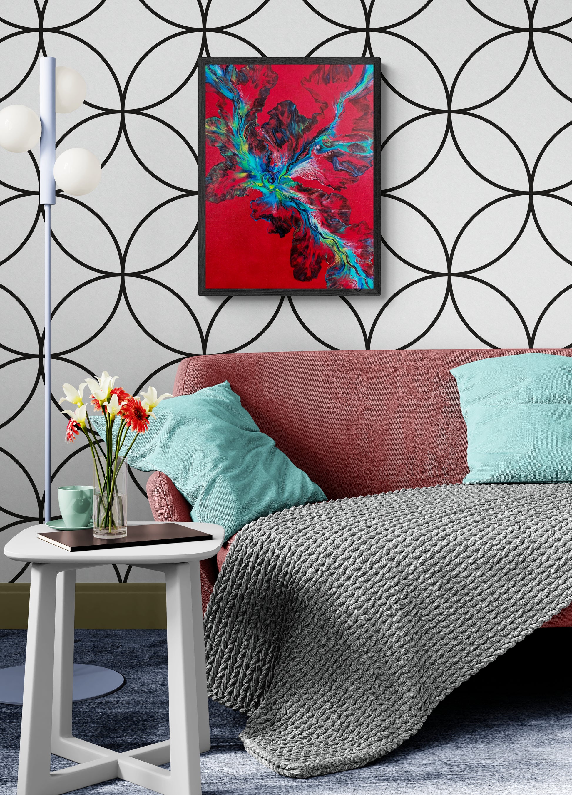 The painting "Elemental Surge" hangs above a coral-pink couch with mint pillows and a chunky gray knit throw. The abstract painting features vivid teal, neon green, and black accents erupting across a red background, set against a white wall with bold black geometric circles.