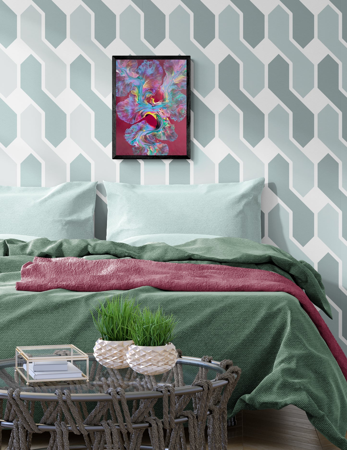 An abstract painting titled "Ember Glow" is framed in black and displayed above a bed with mint green pillows and sage green bedding. The artwork features vibrant pink, teal, gold, and purple hues swirling over a rich burgundy background. The accent wall behind the bed has a white and muted blue hexagonal pattern, while a knotted rope coffee table holds two woven planters with grass, creating a fresh and lively bedroom scene with bold visual contrast.