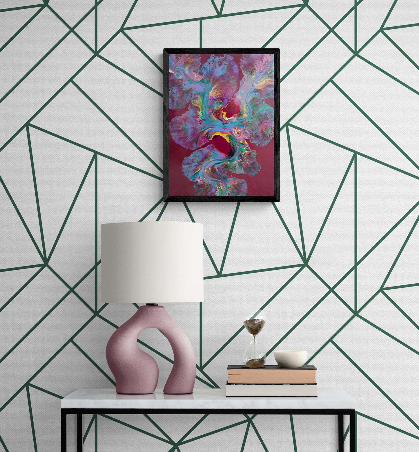 A bold abstract painting titled "Ember Glow" is framed in black and displayed on a white wall with a deep green geometric line pattern. The artwork features radiant teal, pink, purple, and gold hues flowing over a rich burgundy background. Below, a white marble console table holds a mauve sculptural lamp, an hourglass, stacked books, and a small ceramic bowl, creating a modern and artful vignette.