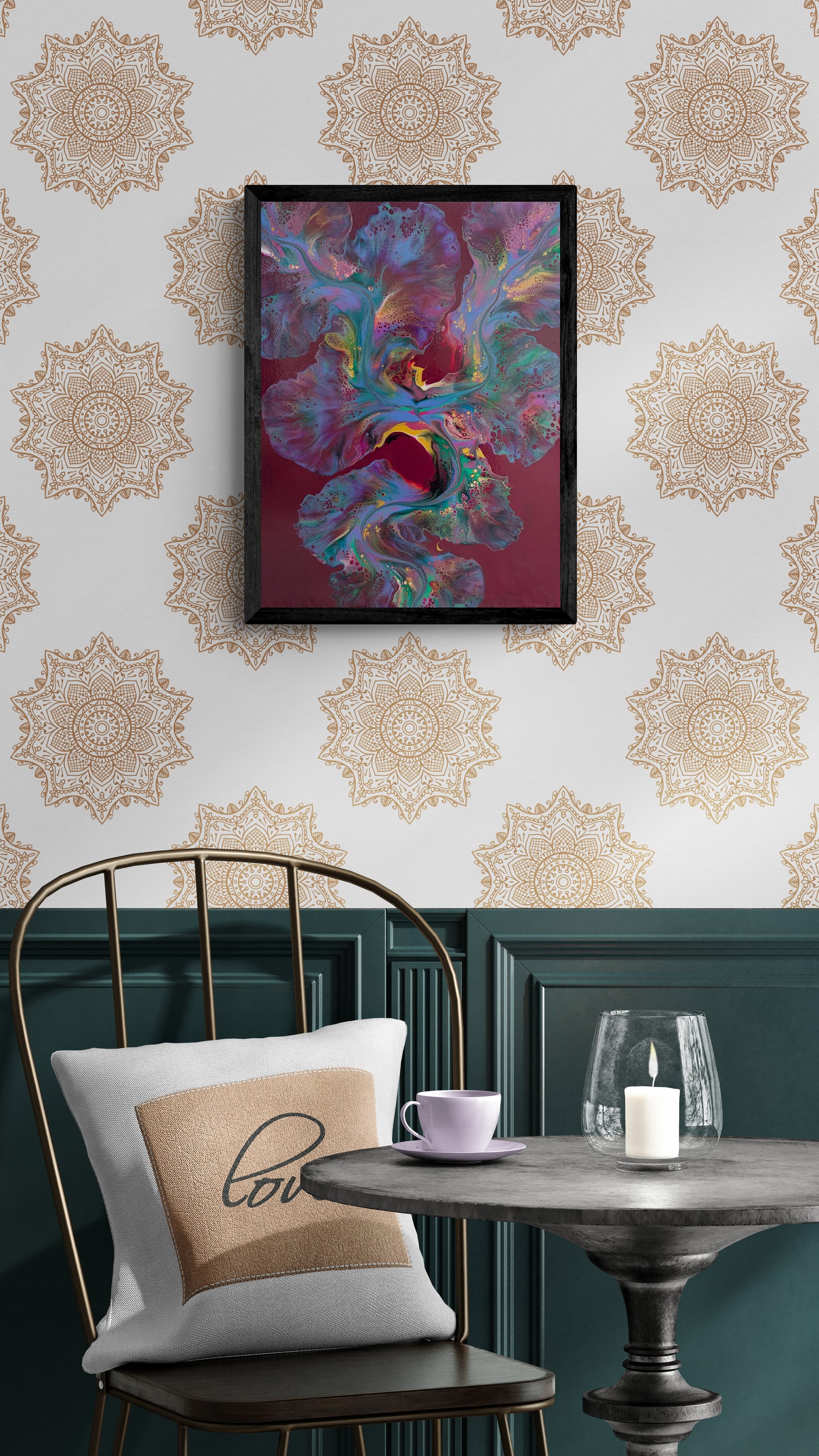 An abstract painting titled "Ember Glow" is framed in black and displayed on a white wall with warm gold mandala patterns. The artwork features fluid shapes in vivid pink, teal, violet, and gold over a deep burgundy background. Below, a cozy seating area includes a black spindle chair with a “love” pillow, a candle, and a lavender cup on a round table, set against dark teal wainscoting for a warm and eclectic ambiance.