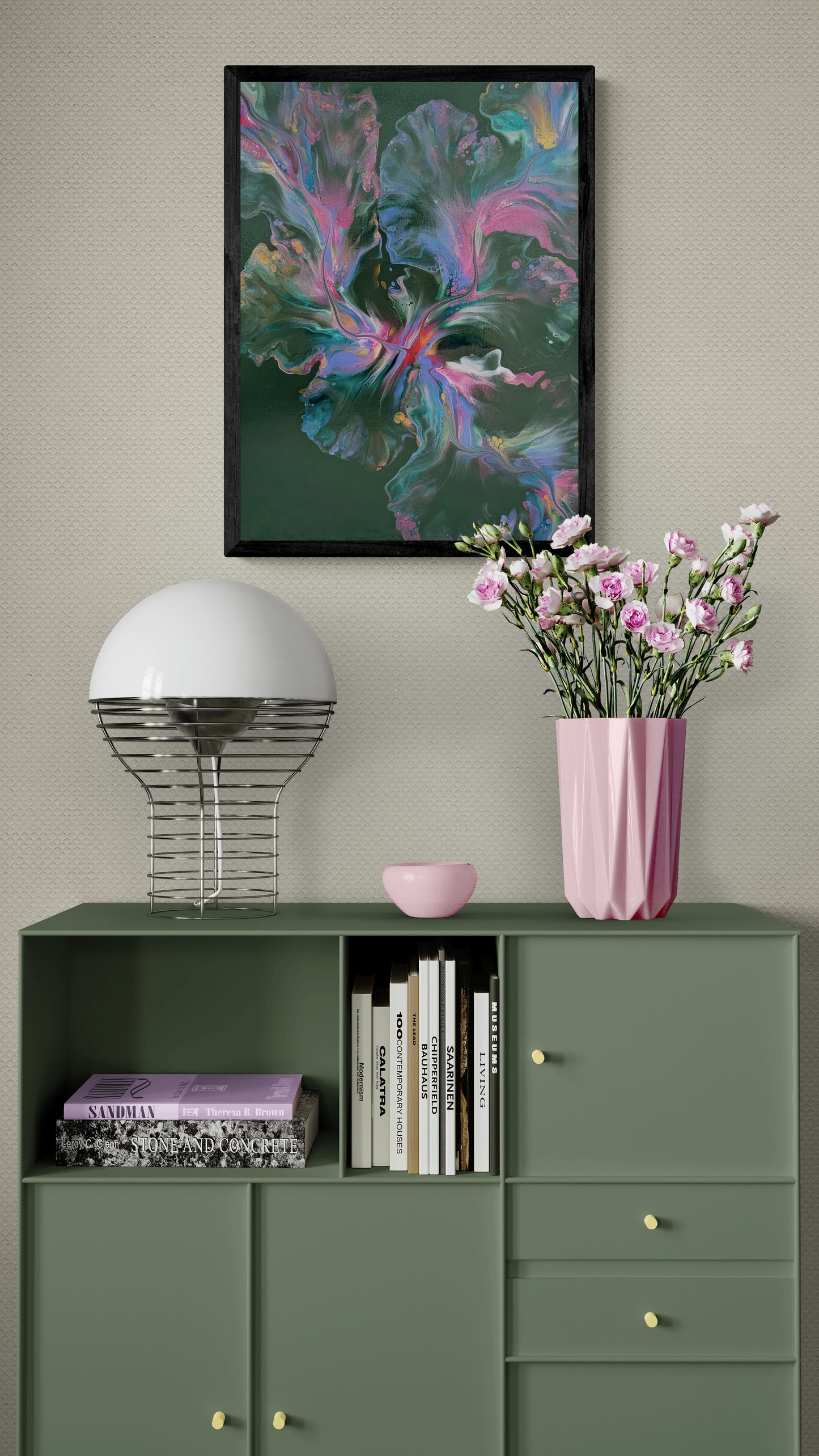 An abstract painting titled "Enchanted Sky" is framed in black and displayed on a soft beige wall above a sage green mid-century modern cabinet. The artwork features flowing strokes of pink, teal, lavender, and gold over a deep green background. The cabinet is styled with a modern chrome lamp, a stack of books, and a tall pink vase with blush flowers, creating a calm, artistic interior with soft color echoes and minimalist charm.
