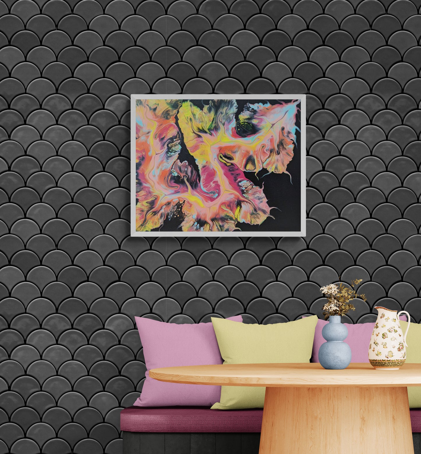 Framed abstract painting with bold pink, yellow, green, and black fluid art displayed on a scalloped black tile wall above a cozy nook. The nook features a round wooden table, pastel pillows in lavender and light green, and soft-toned ceramic vases with dried flowers.