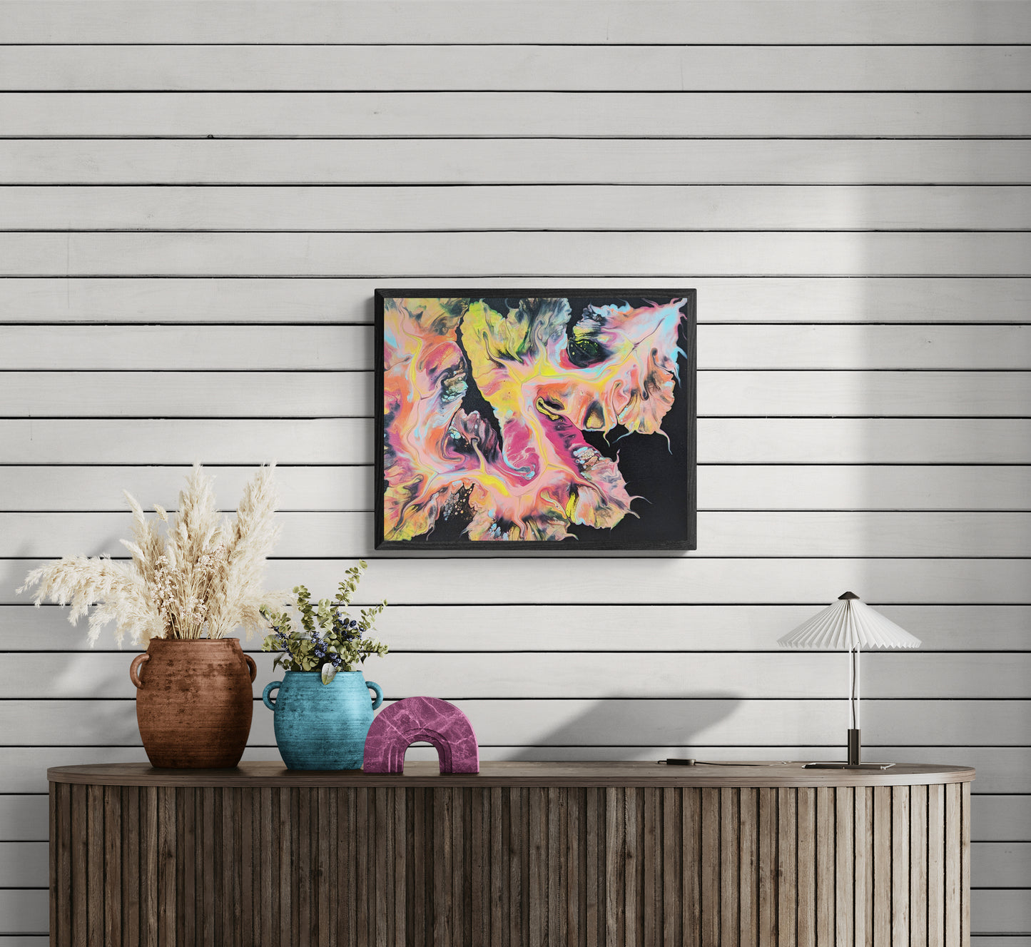 Framed abstract painting titled "Festival Dragon" hung on a white shiplap wall above a wood sideboard with decorative vases and a table lamp. The painting features vivid swirls of yellow, pink, and teal on a black background.