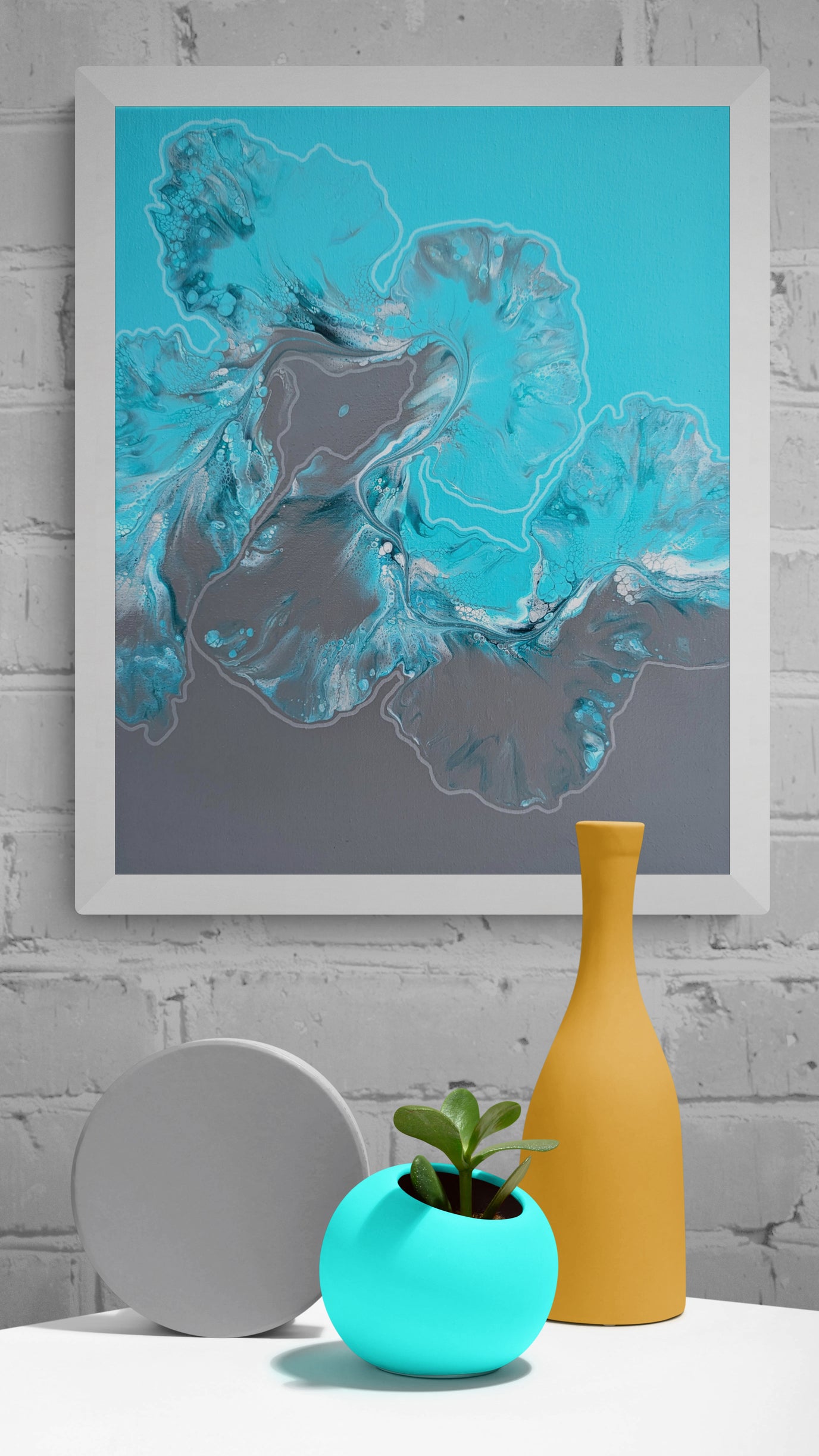 Framed fluid acrylic painting in icy turquoise and gray tones with delicate white outlines, hanging on a white brick wall above a white table with sculptural decor including a turquoise planter with a succulent, a tall yellow vase, and a round gray object.