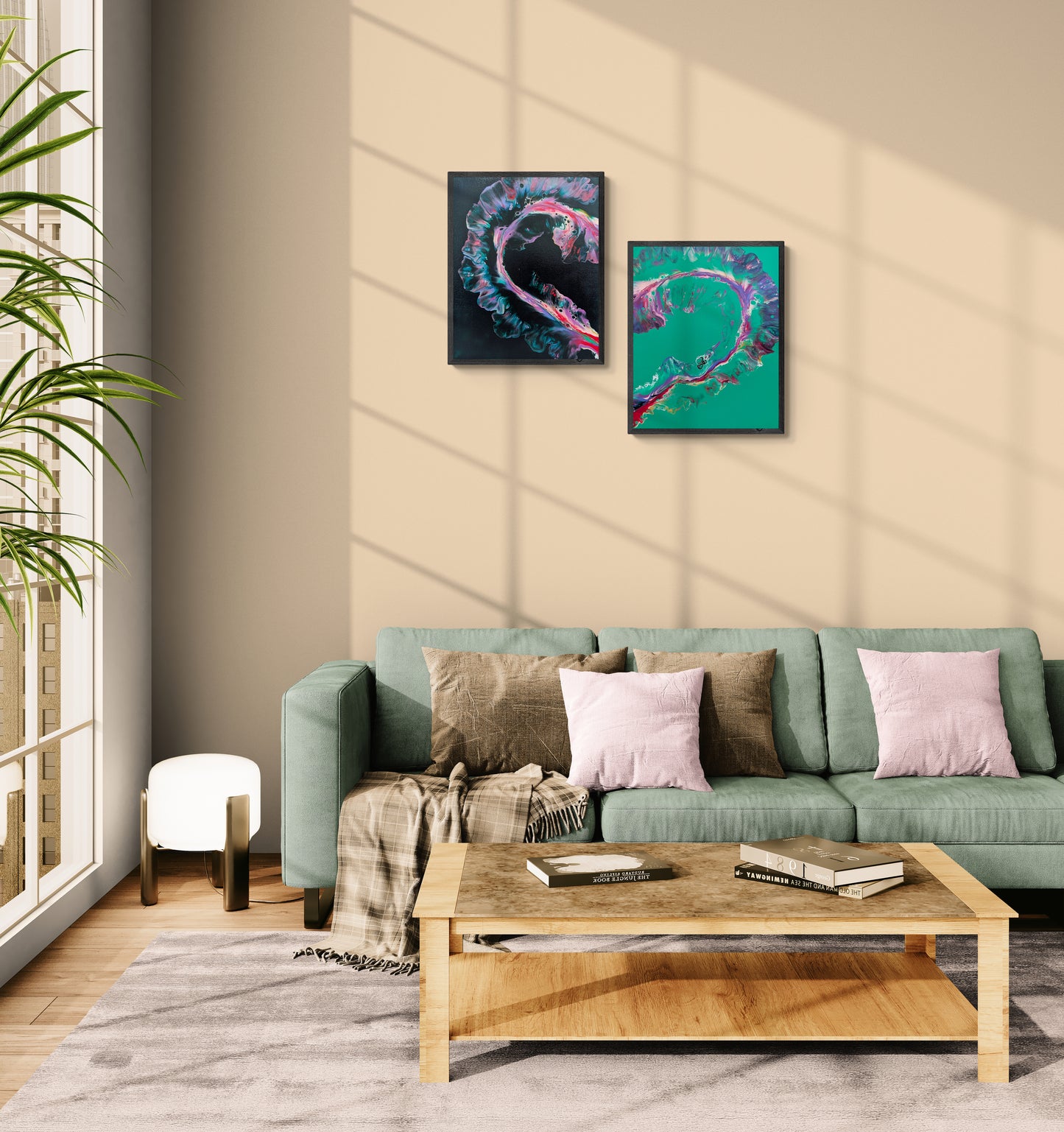 Two abstract fluid paintings forming a heart shape are displayed one above the other on a beige wall in a sunny living room. One painting has a black background, the other a green one, both with vivid swirling colors in pink, purple, teal, and red. Below the artwork is a green sofa with pink and neutral pillows, a soft gray rug, and a wood coffee table. Natural light casts soft shadows across the scene.