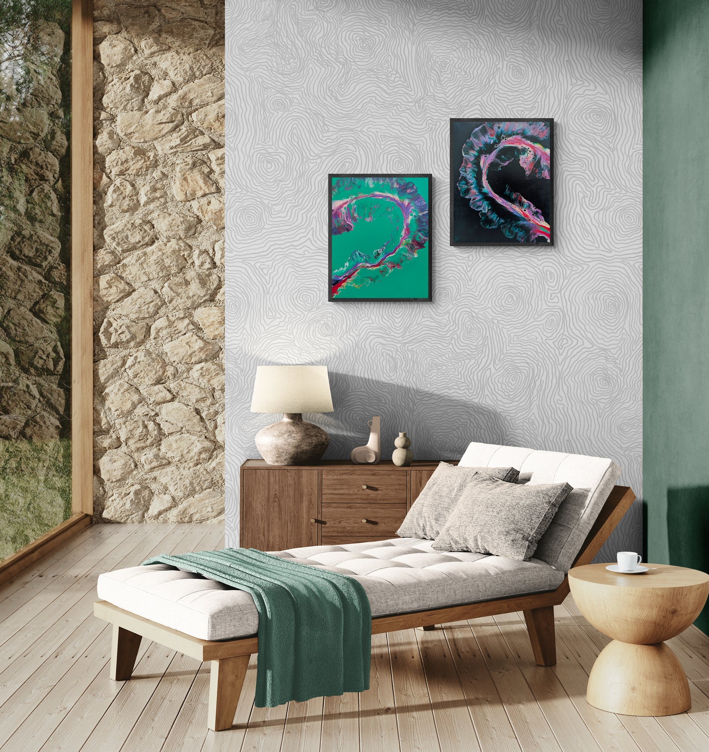 Two abstract paintings forming a colorful heart are displayed on a textured gray wall above a modern wood sideboard. The left canvas features a green background, the right a black one, both with swirling pink, teal, and purple accents. The room includes a wood-framed chaise lounge with a light cushion, a green throw blanket, and a round wood side table. A stone wall and floor-to-ceiling window bring natural light and organic warmth to the scene.