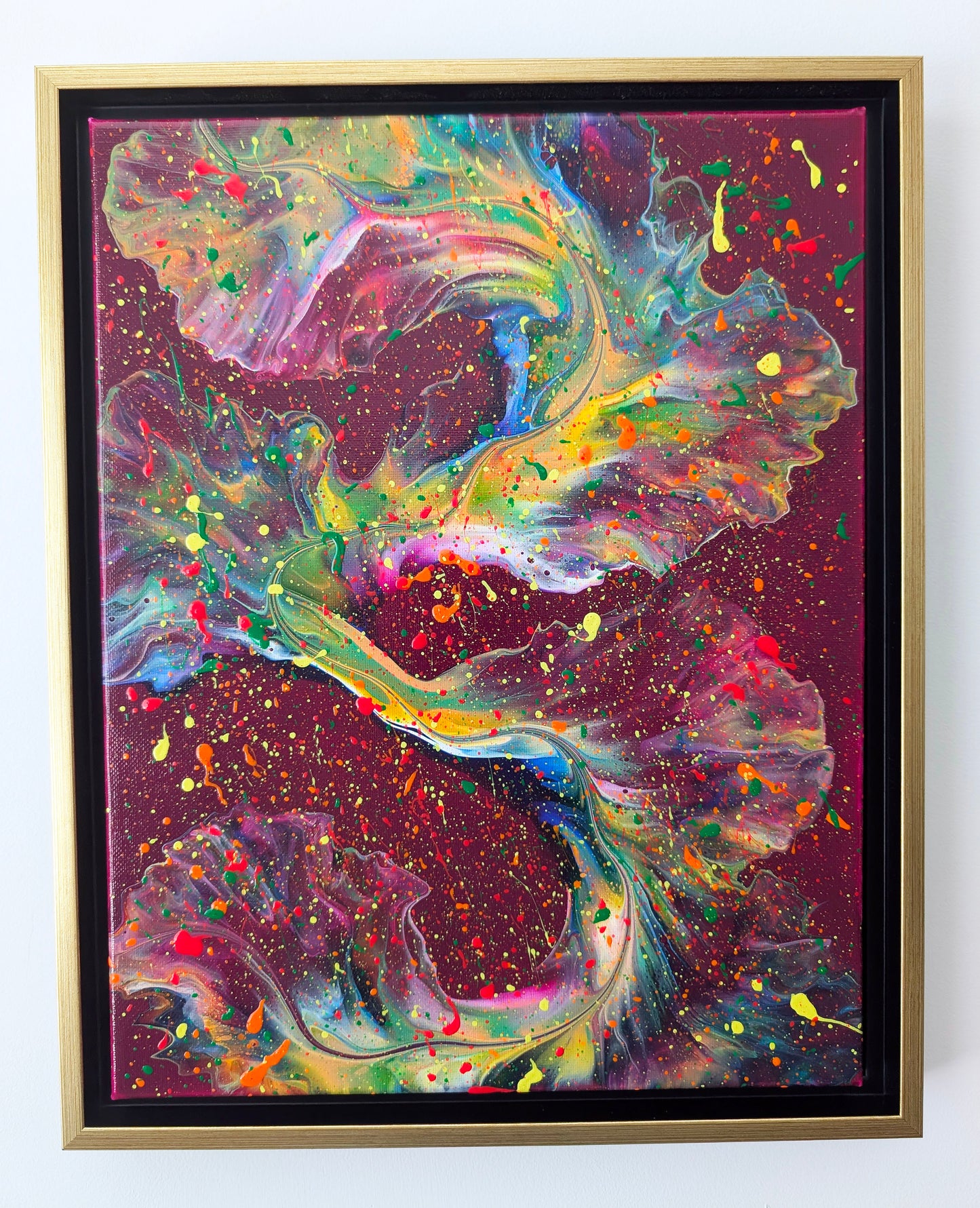 Original abstract fluid art painting titled 'I Wish I Had Your Confidence,' featuring bold ribbons of neon green, yellow, orange, and pink swirling across a dark magenta background, layered with splashes of bright red, green, and yellow. Displayed in a gold floating frame against a white wall.