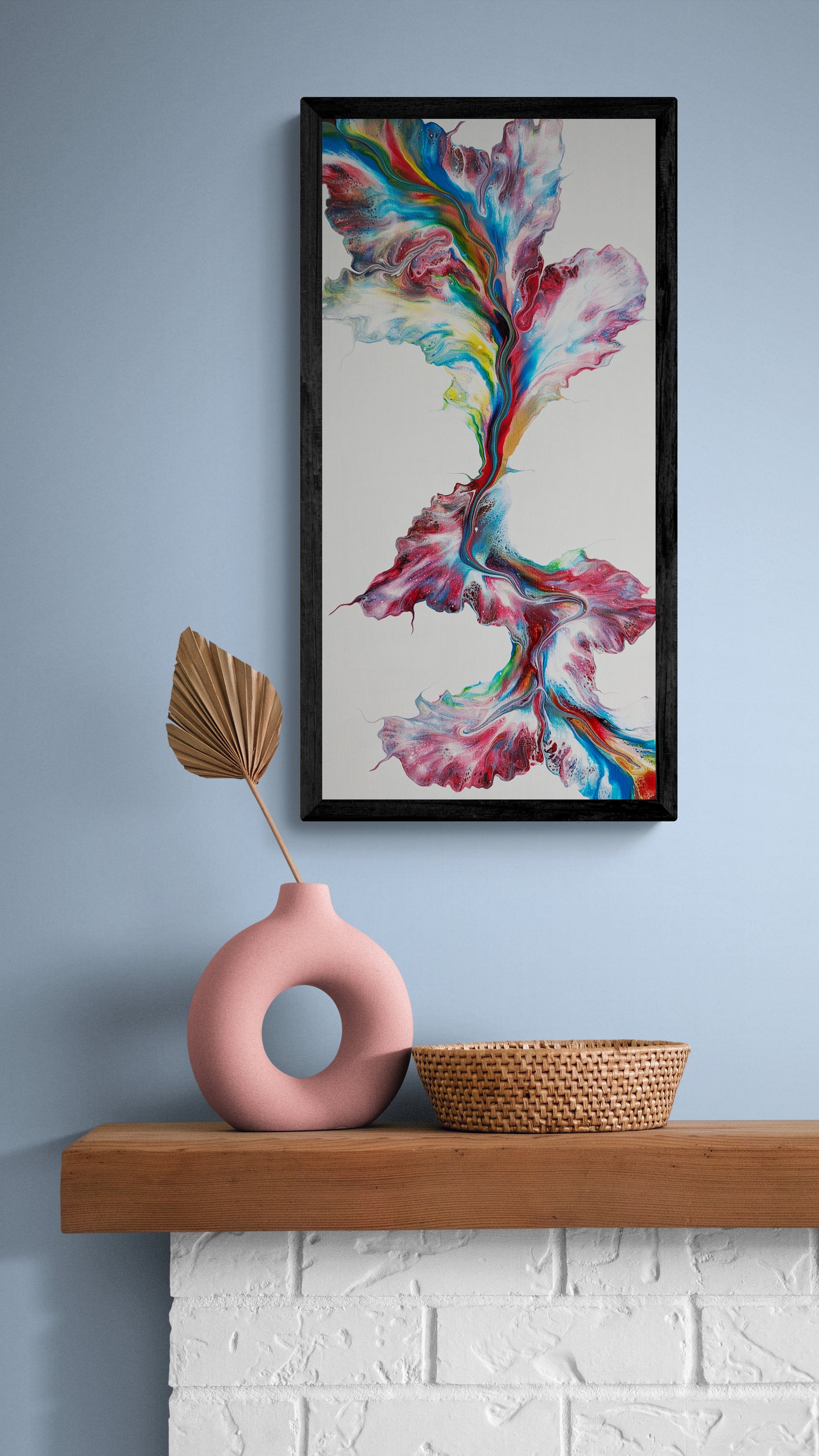 A bold abstract painting titled "Macaw" is framed in black and hung on a dusty blue wall above a rustic wood mantle. The artwork features vivid red, blue, yellow, and green tones flowing vertically over a clean white background. Below the painting sits a round pink sculptural vase holding a dried palm leaf, next to a woven basket, all resting on a wooden shelf with a white painted brick base, creating a modern-yet-earthy styled vignette.