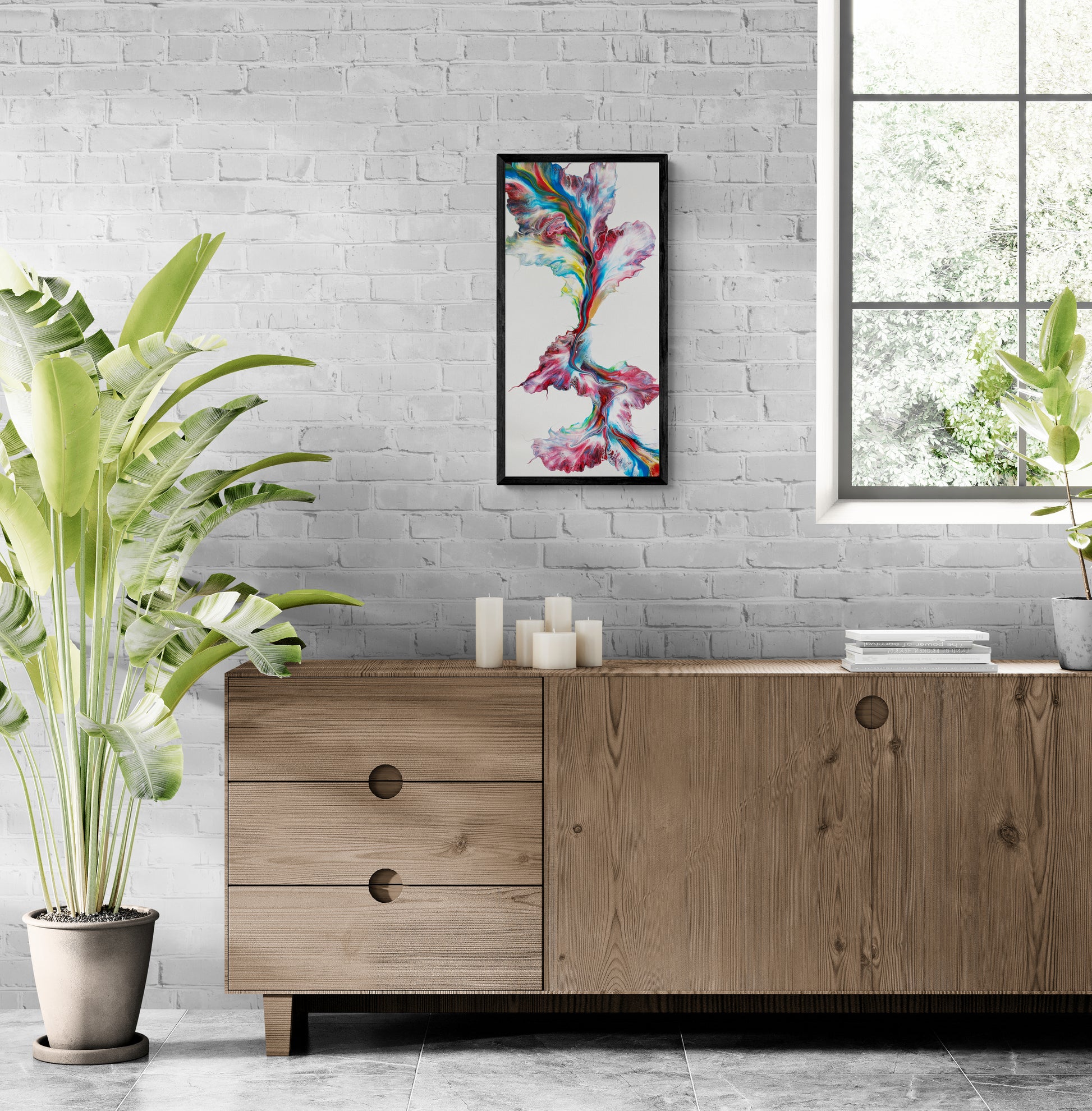 A vibrant abstract painting titled "Macaw" is framed in black and displayed on a whitewashed brick wall above a light wood sideboard. The artwork features a vertical ribbon of swirling red, teal, yellow, and cobalt hues over a white background, evoking the energy of feathers in flight. The room is styled with stacked books, minimalist white candles, a large tropical plant in a gray pot, and a window letting in soft natural light, creating a serene and stylish living space.