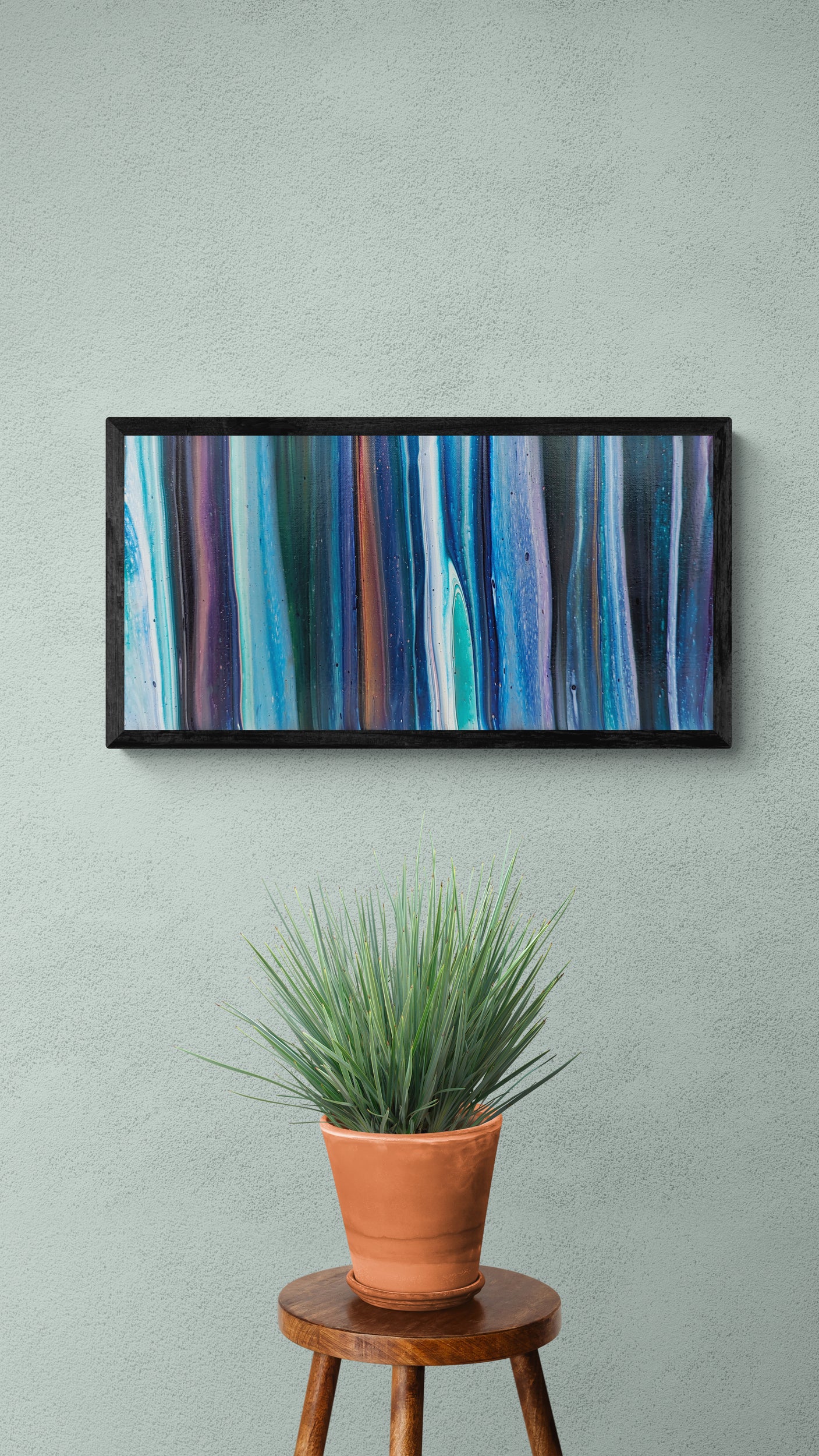 Framed abstract fluid painting with vertical flows of blue, turquoise, violet, and gold, displayed on a pale sage wall above a terracotta potted plant on a wooden stool.