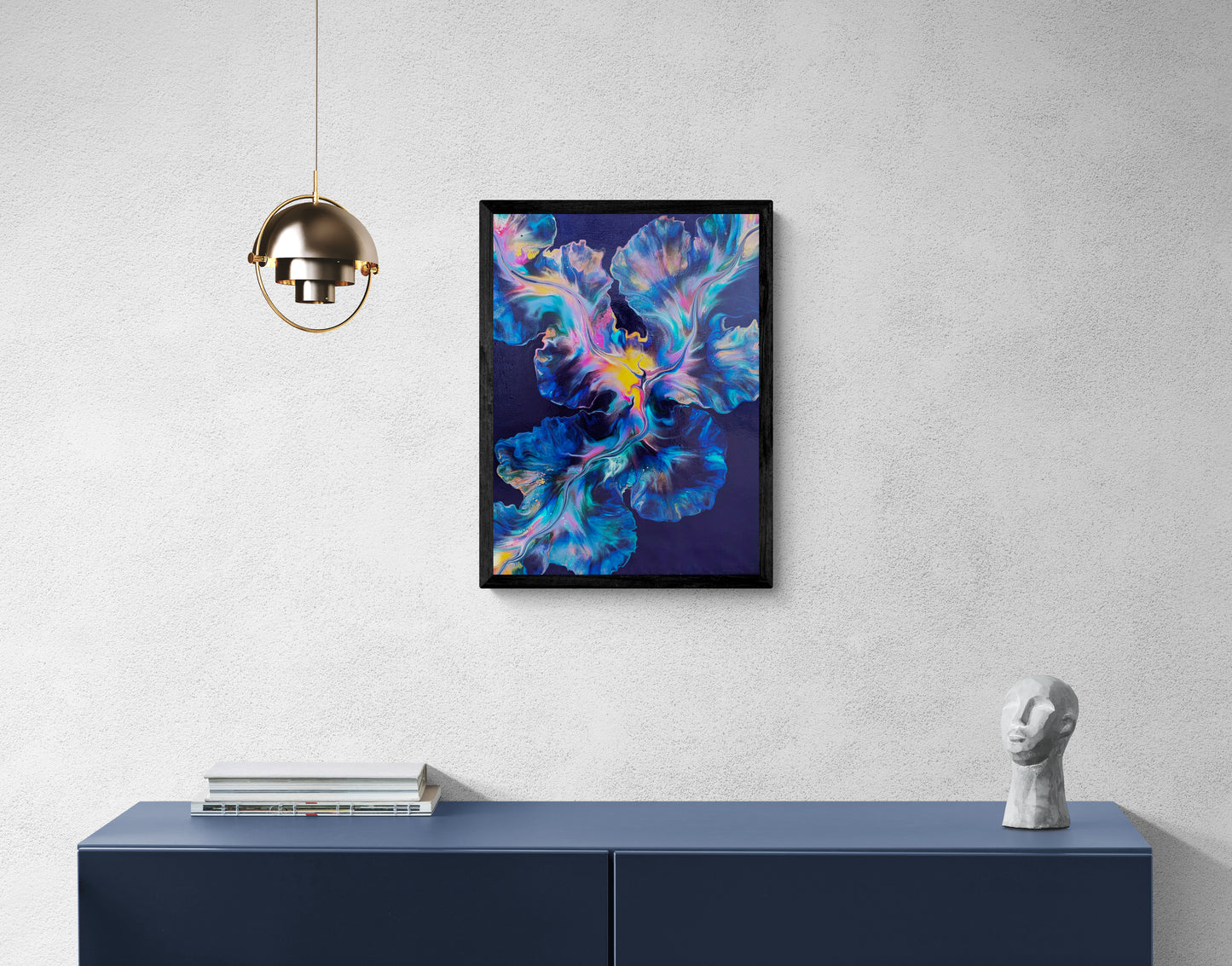 An abstract painting titled "Nebula Bloom" is framed in black and displayed on a light gray wall above a navy blue credenza. The artwork features radiant blends of bright pink, cobalt, gold, and turquoise swirling from a glowing yellow center over a deep navy background. The scene includes a sculptural white bust, a stack of books, and a gold hanging pendant light, creating a modern, minimalist interior that highlights the painting’s cosmic energy.