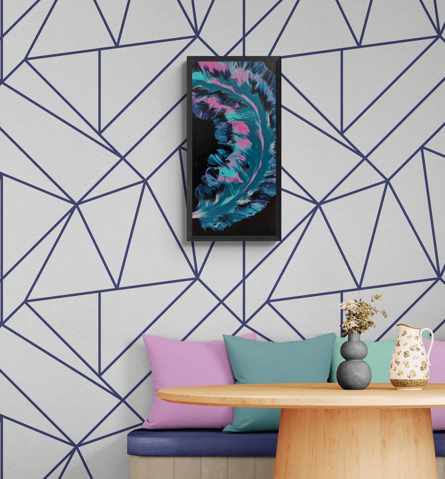 Framed abstract painting with swirling teal, pink, and deep purple tones on a black background, displayed above a pastel-accented seating nook with geometric wallpaper and a round wooden table with a ceramic pitcher and vase.