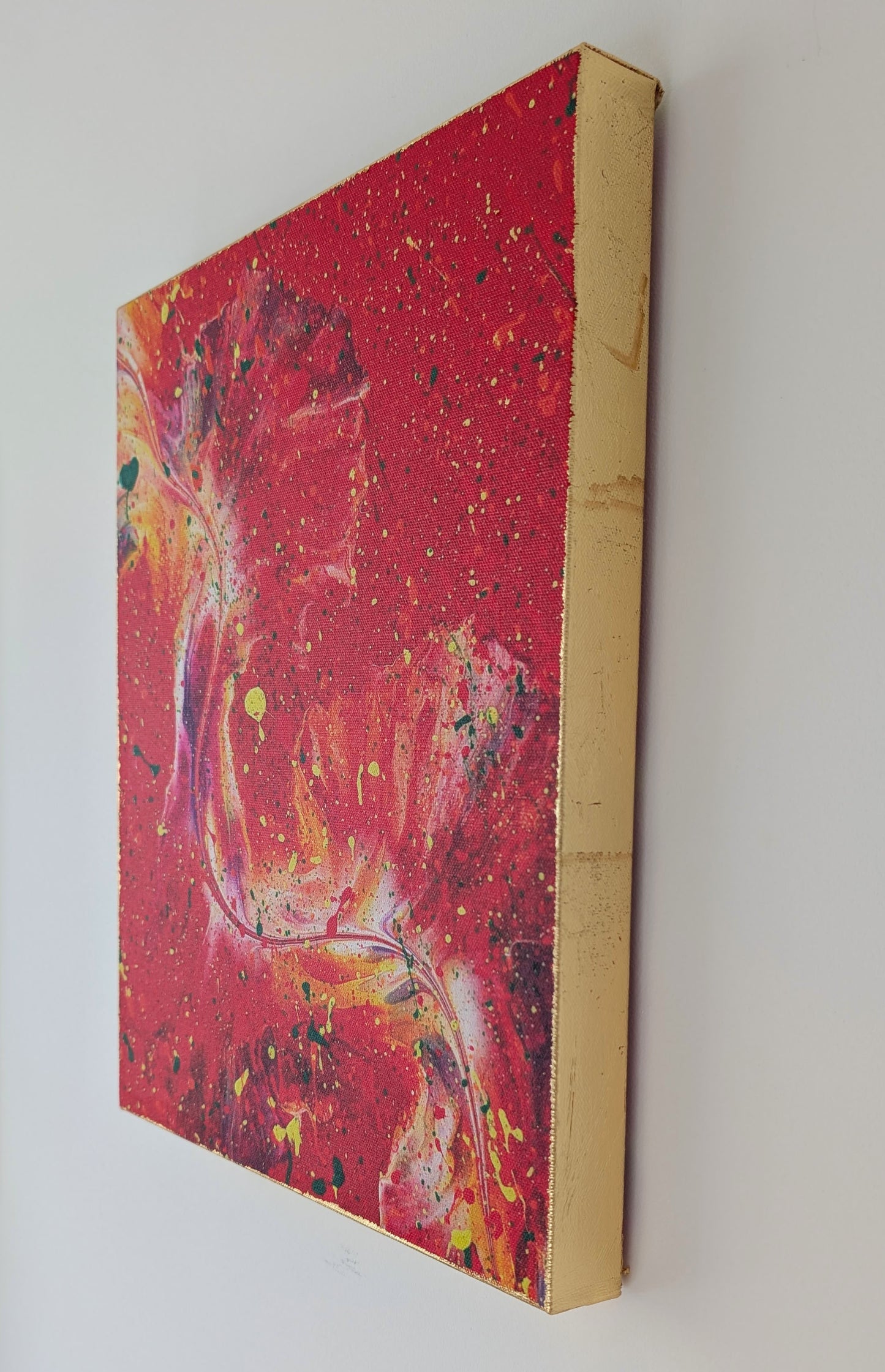 Close side view of the red fluid art canvas hanging on a wall, highlighting the gold leaf texture on the edges and the bold layered paint splatters on the front.