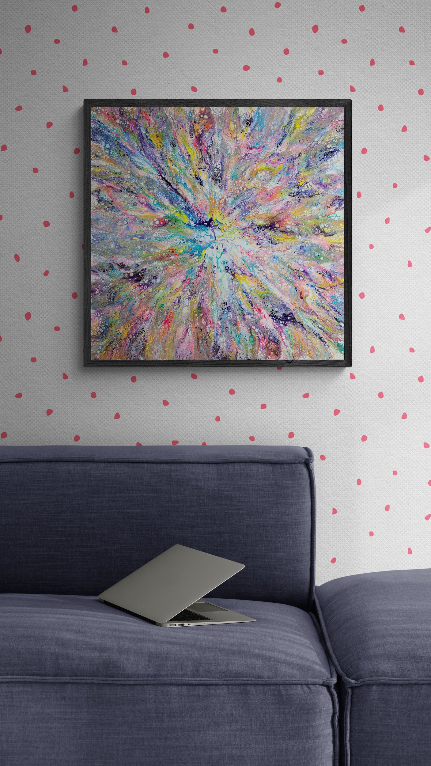 A square abstract fluid art painting with a radiant burst of rainbow colors is framed and hung on a light gray wall with soft pink polka dots. Below the painting sits a deep blue couch with a slightly open laptop resting on the cushion, creating a relaxed and creative atmosphere.