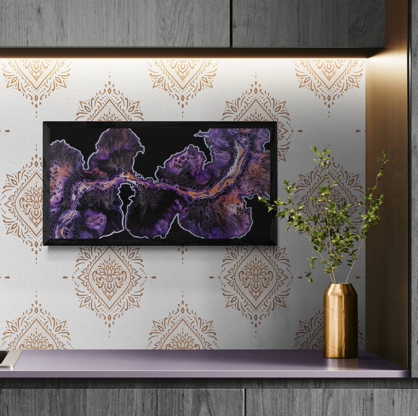 Framed abstract painting in deep purple, and copper tones displayed above a soft lavender counter against a patterned gold accent wall, with a gold vase holding greenery beside it.