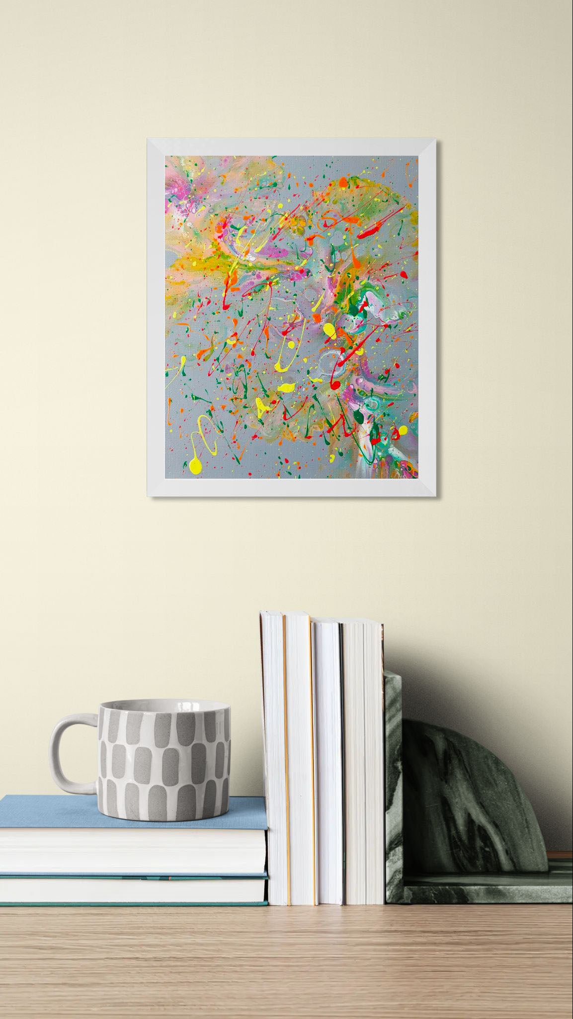 Framed colorful abstract painting featuring pink, yellow, green, and red splatters on a gray canvas, hung above a stack of books and a gray-patterned mug.