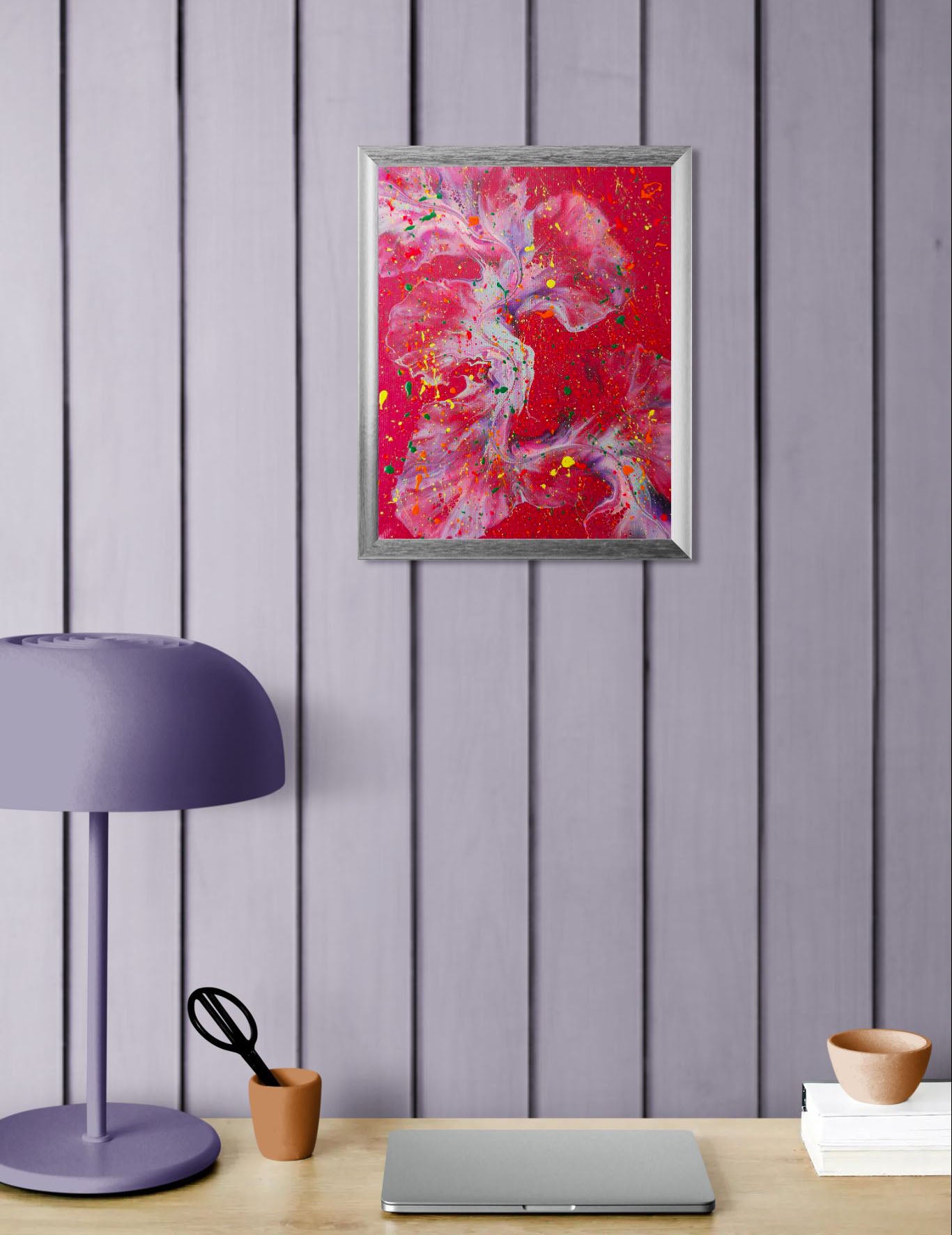 A bold, red-based abstract painting with dynamic swirls of purple and blue, highlighted by vivid neon splatter in yellow, pink, orange, and green. The painting is framed in silver and hangs above a workspace with a lavender lamp, black scissors in a clay holder, and a closed laptop