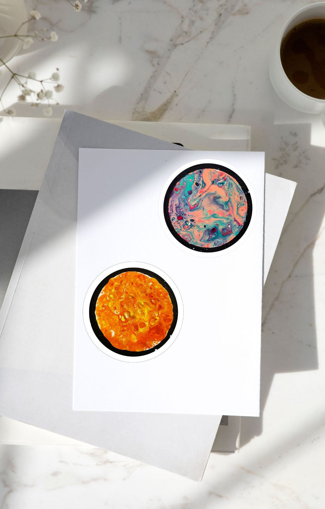 Two glossy moon-themed stickers—one in swirling pastel pinks and purples, the other in fiery oranges—applied to the cover of a white journal, placed on a marble surface next to a cup of coffee.