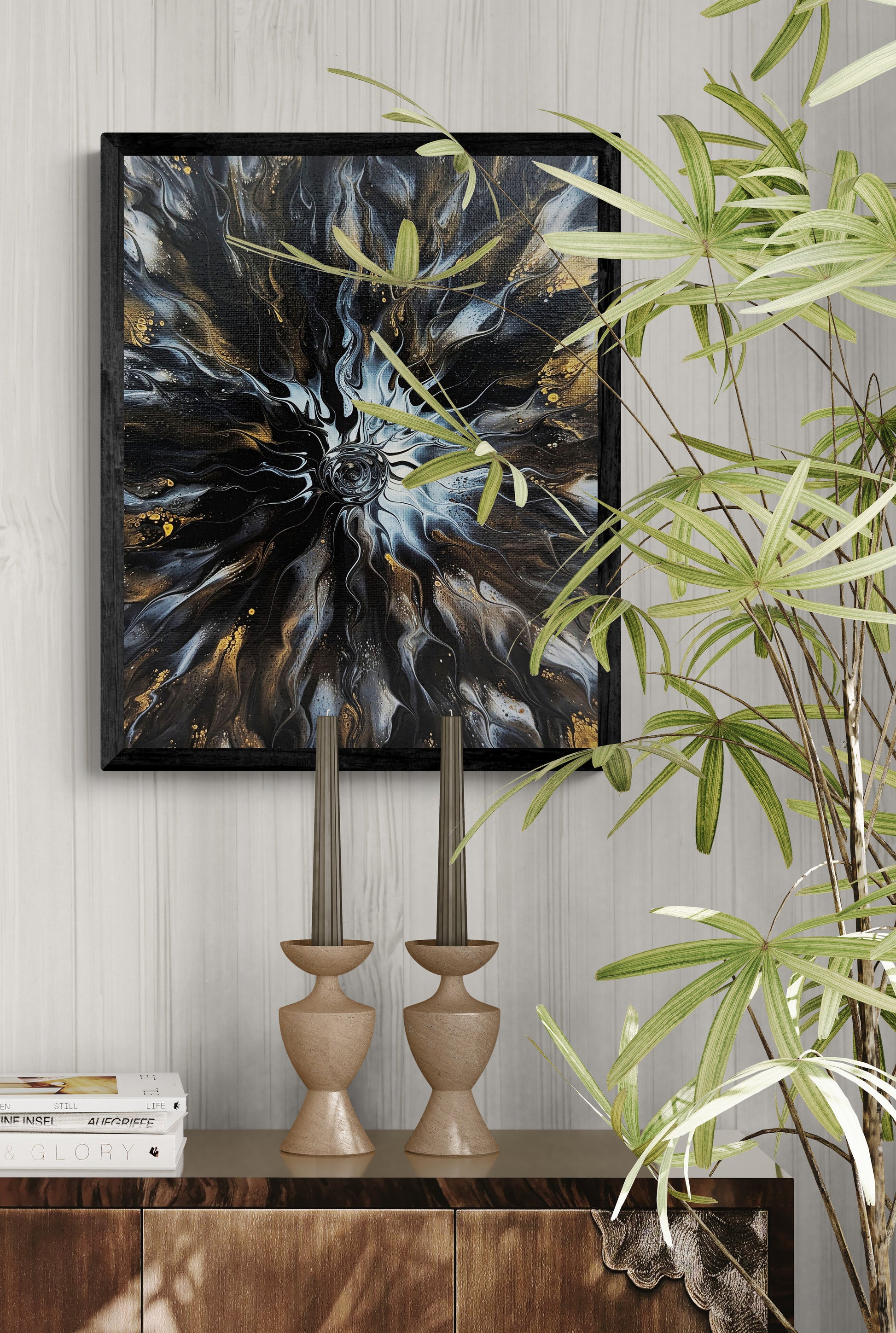 Black-framed abstract fluid art print with flowing gold and gray tones hanging above a wooden cabinet with candlesticks, books, and green plants