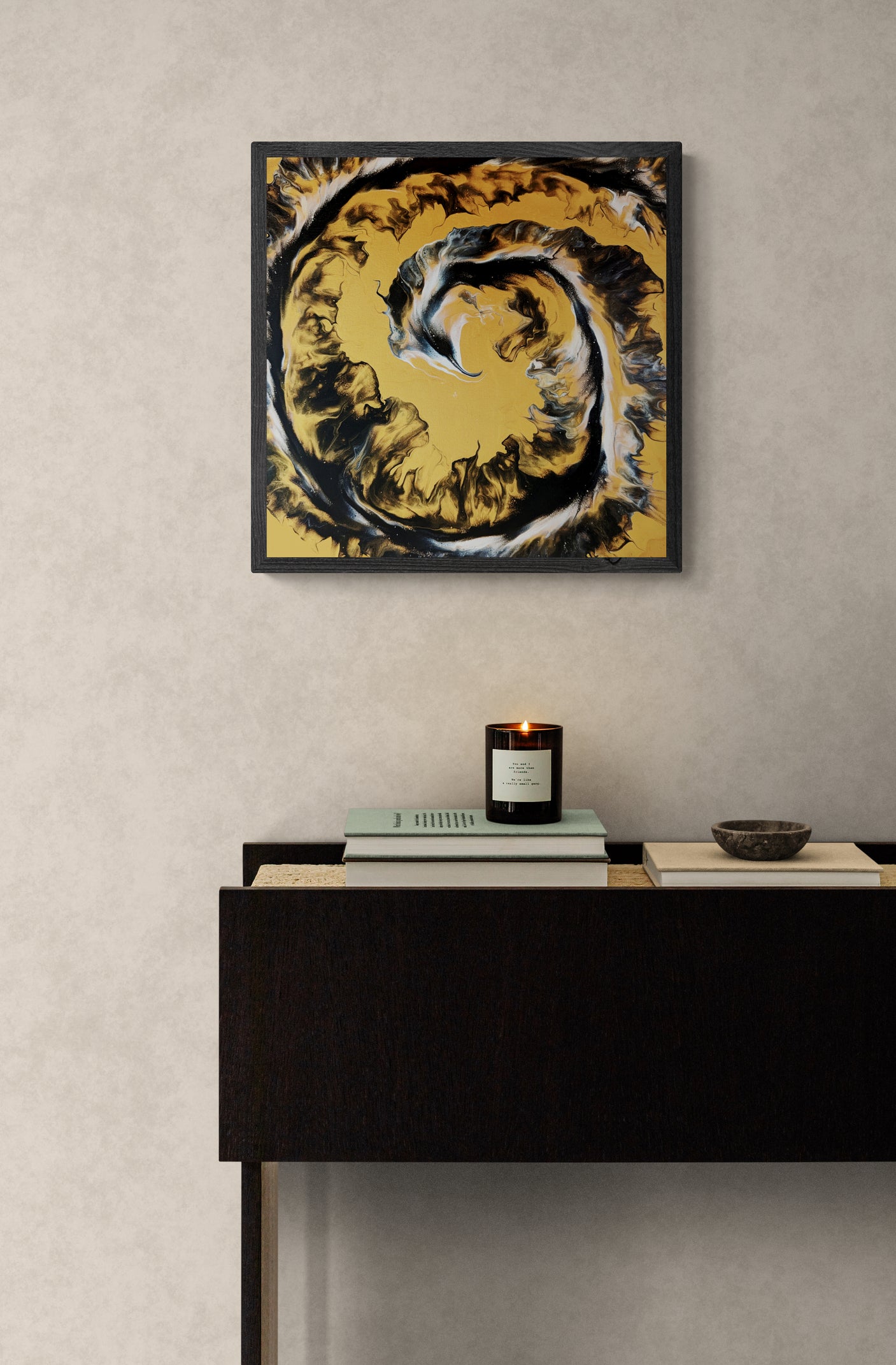 A square abstract painting in golden ochre, black, and white with a spiraling, smoke-like pattern hangs on a soft beige textured wall. Below it sits a dark wood console table with a lit black candle, stacked books, and a small ceramic bowl, creating a cozy, minimalist interior with earthy elegance.