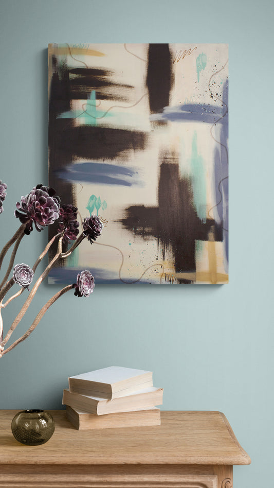 A vertical abstract painting from the "Between the Layers" series displayed on a soft blue wall. The composition includes vertical blocks of raw umber, splashes of teal and lavender, and delicate dark splatters. In the foreground, a stack of books and a small glass vase sit on a light wood console.