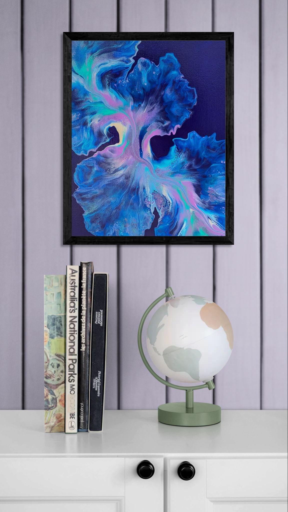 Framed abstract fluid painting in cool blue, purple, teal, and pink tones, styled on a lavender paneled wall above a white cabinet with a sage globe and stacked books.