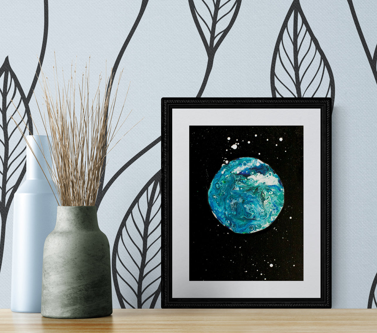 Blue Moon art print in a black frame displayed on a wood surface with modern vases and leaf-patterned wallpaper