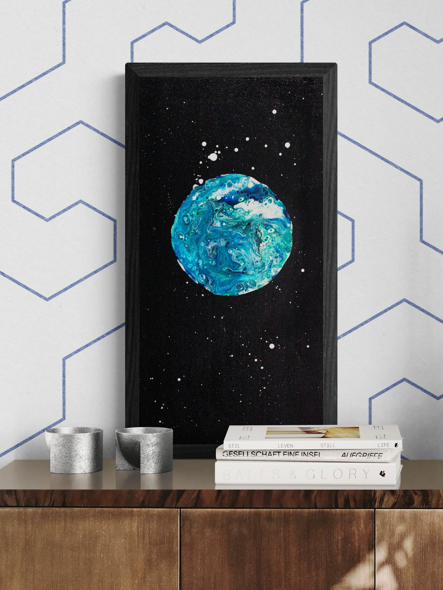 Framed Blue Moon painting showcased above a wooden cabinet styled with sculptural silver decor and modern books, against a white wall with blue-lined patterns.