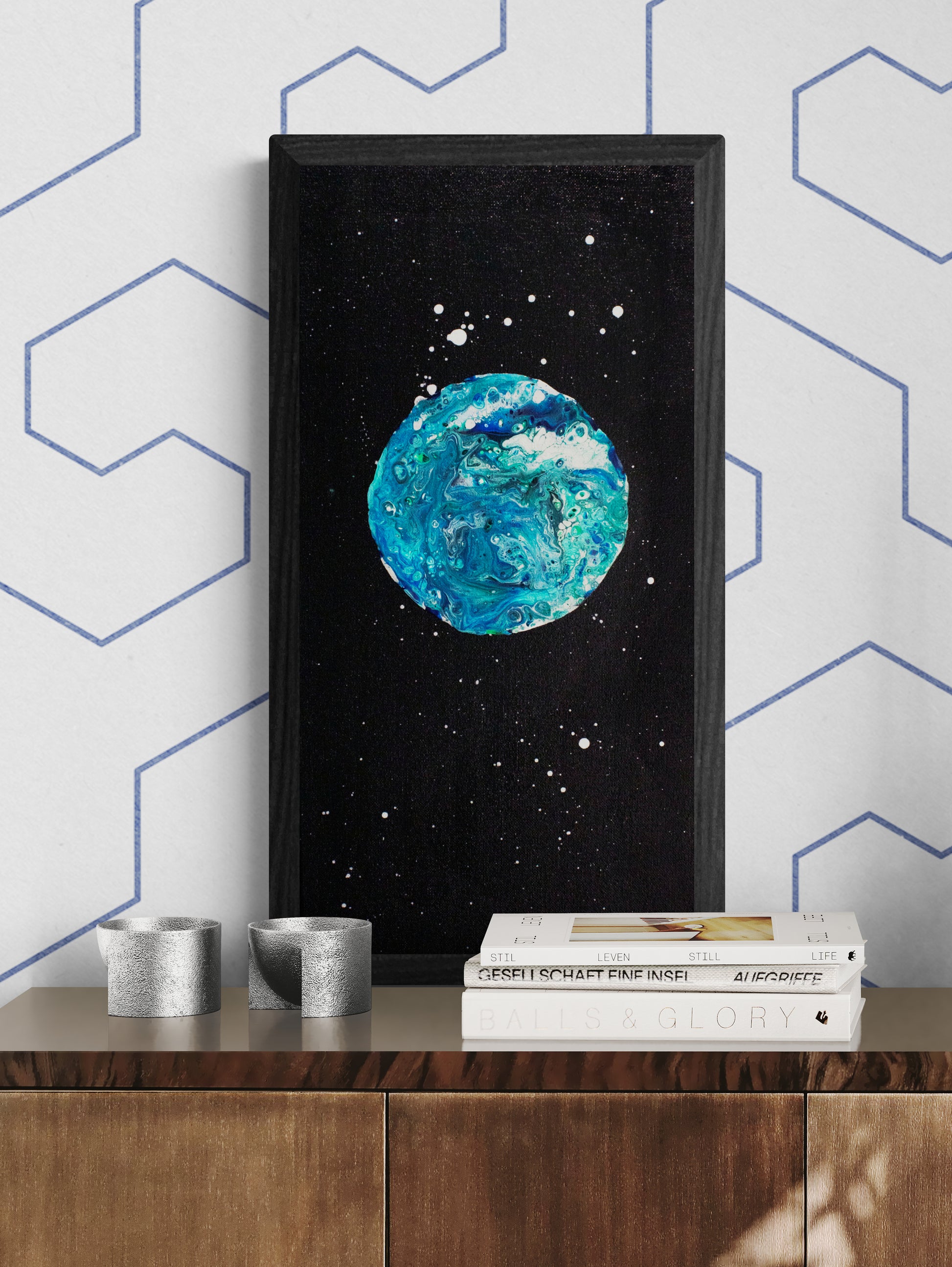 Framed Blue Moon painting showcased above a wooden cabinet styled with sculptural silver decor and modern books, against a white wall with blue-lined patterns.