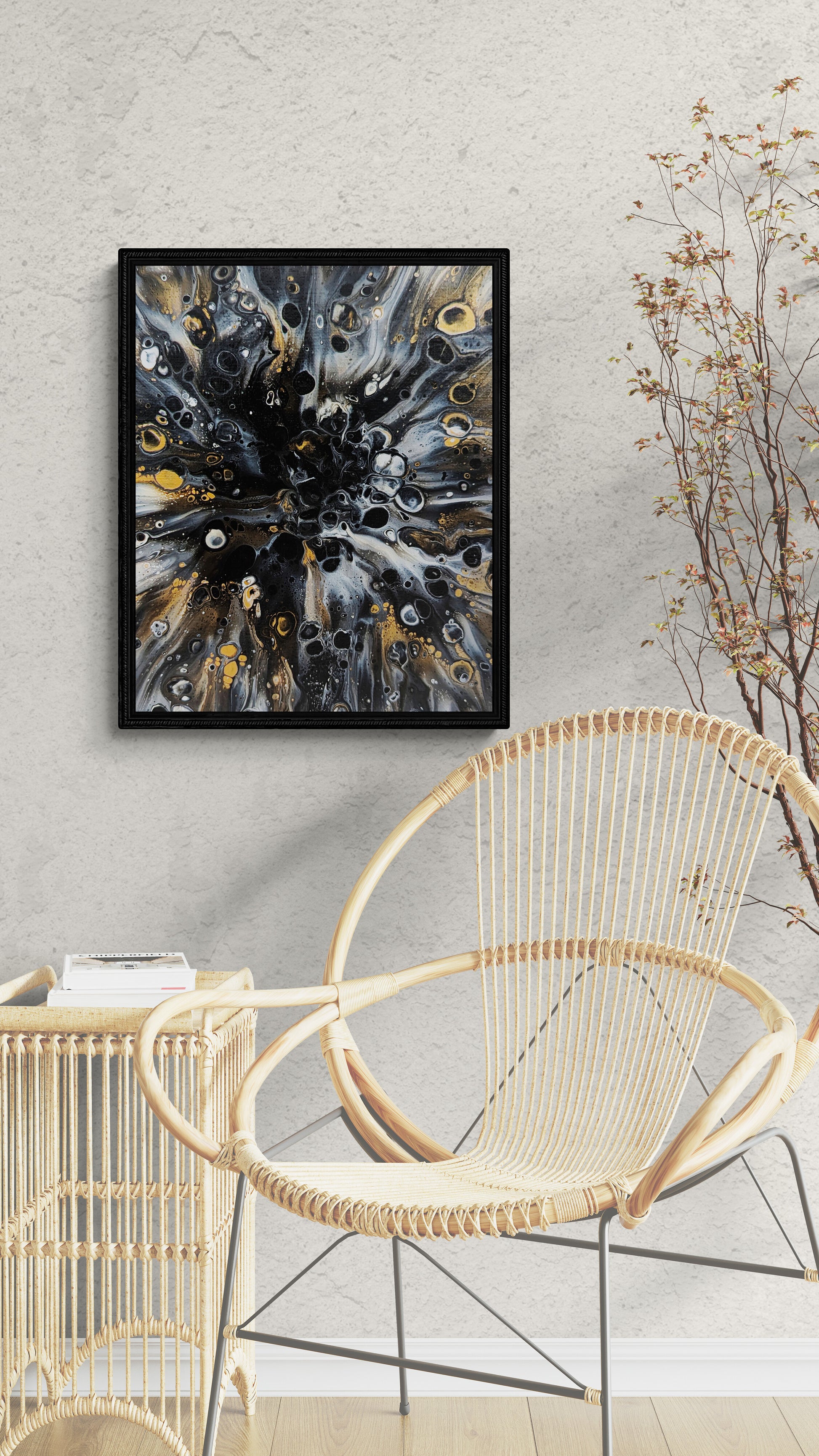 Black-framed abstract fluid art print in black, gold, and gray tones displayed above a rattan chair and side table with a minimalist modern vibe.