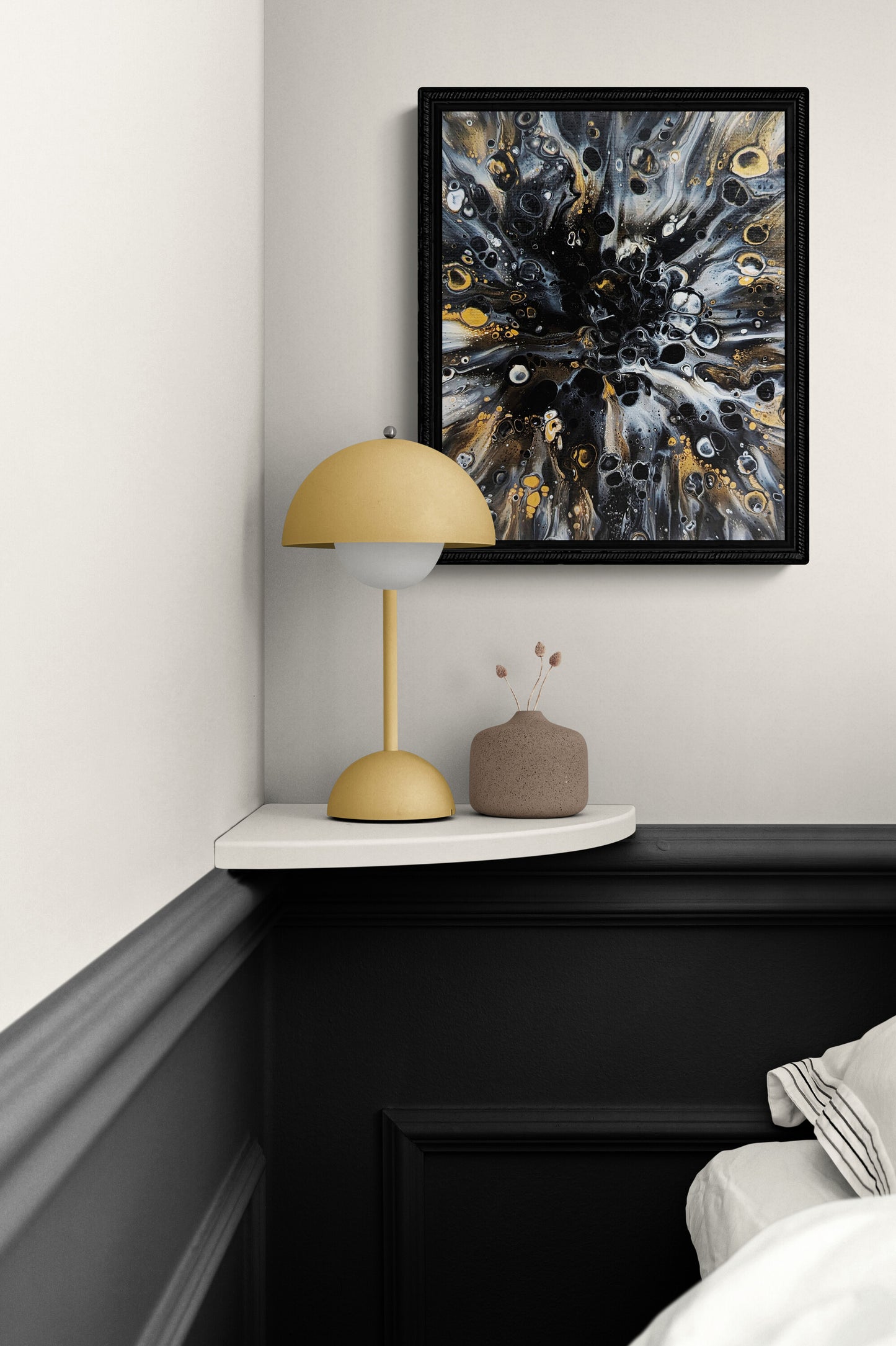 Abstract fluid art print with black frame and gold accents hanging above a small corner shelf with a mustard lamp and neutral vase