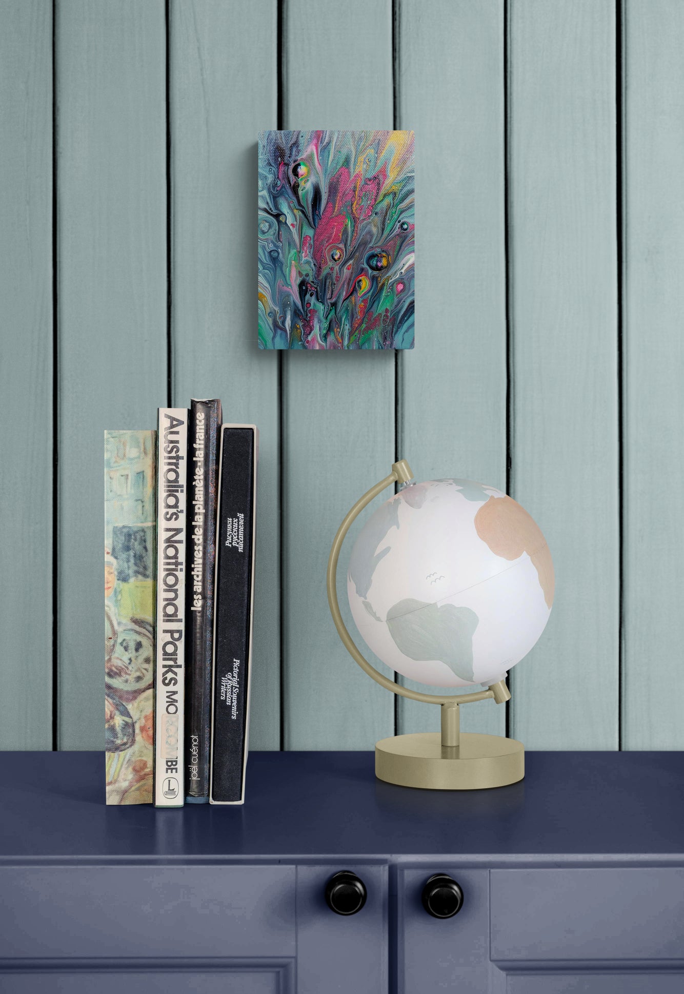 Cheeky, a colorful 5x7 inch acrylic fluid painting, hangs above a navy dresser with a globe and stacked books. The art features electric pinks, teal, and golden yellow with bold cell textures