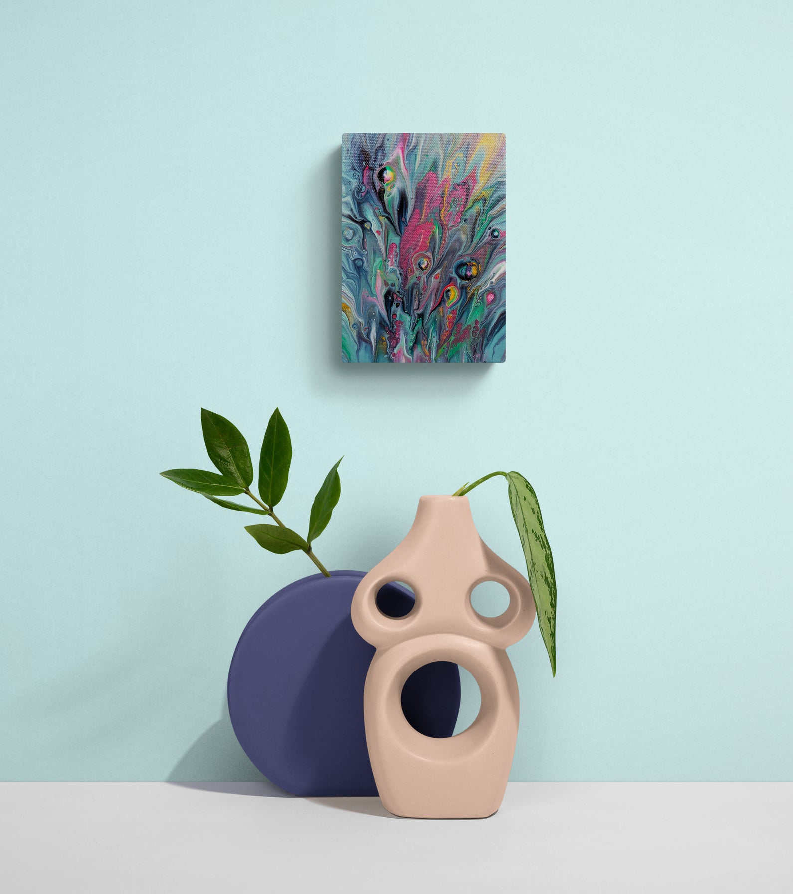 Cheeky painting on a pastel blue wall above a playful arrangement of sculptural beige and navy vases with green foliage. The vivid acrylic artwork features bursts of pink, turquoise, and yellow.