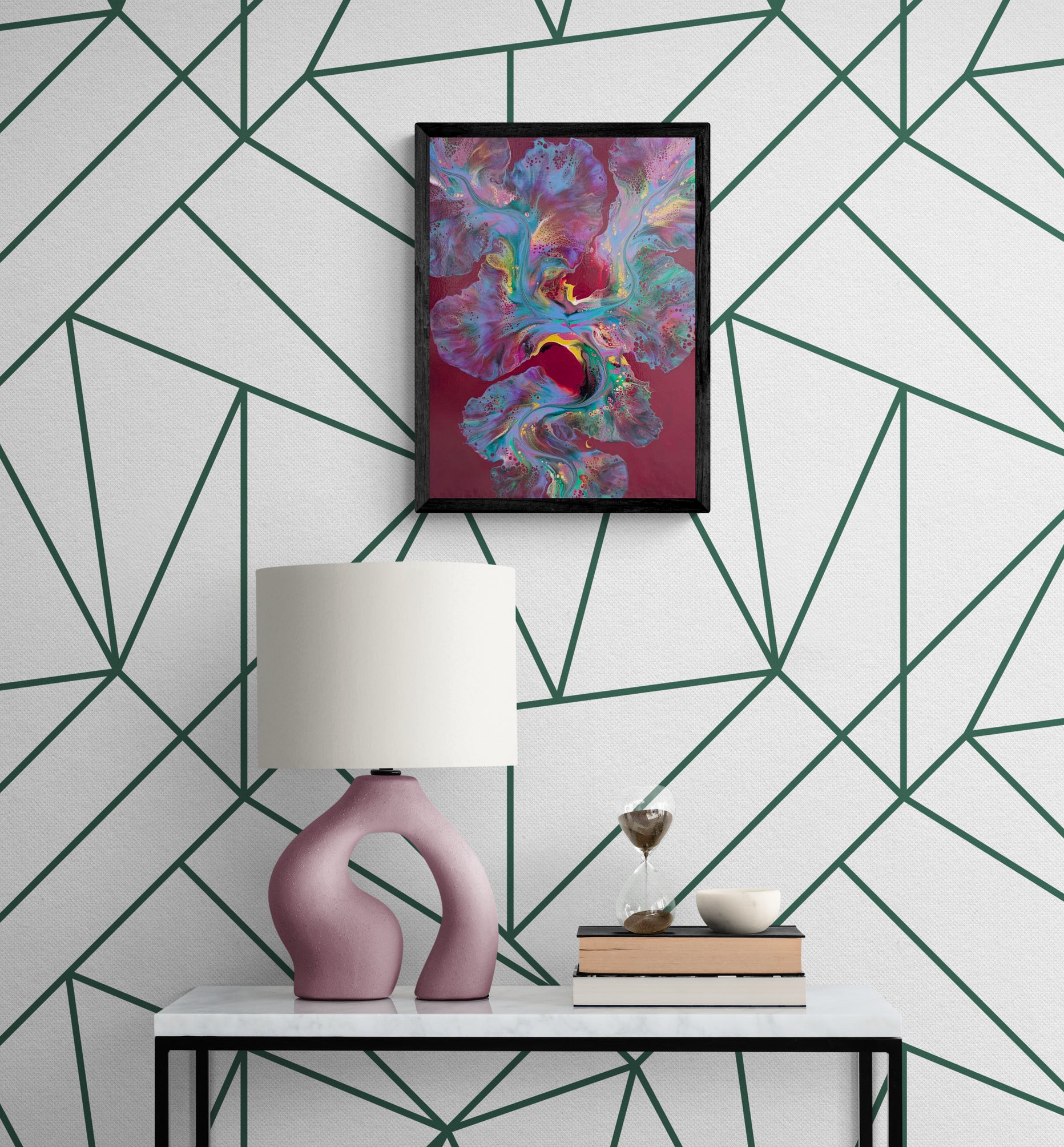 A bold abstract painting titled "Ember Glow" is framed in black and displayed on a white wall with a deep green geometric line pattern. The artwork features radiant teal, pink, purple, and gold hues flowing over a rich burgundy background. Below, a white marble console table holds a mauve sculptural lamp, an hourglass, stacked books, and a small ceramic bowl, creating a modern and artful vignette.