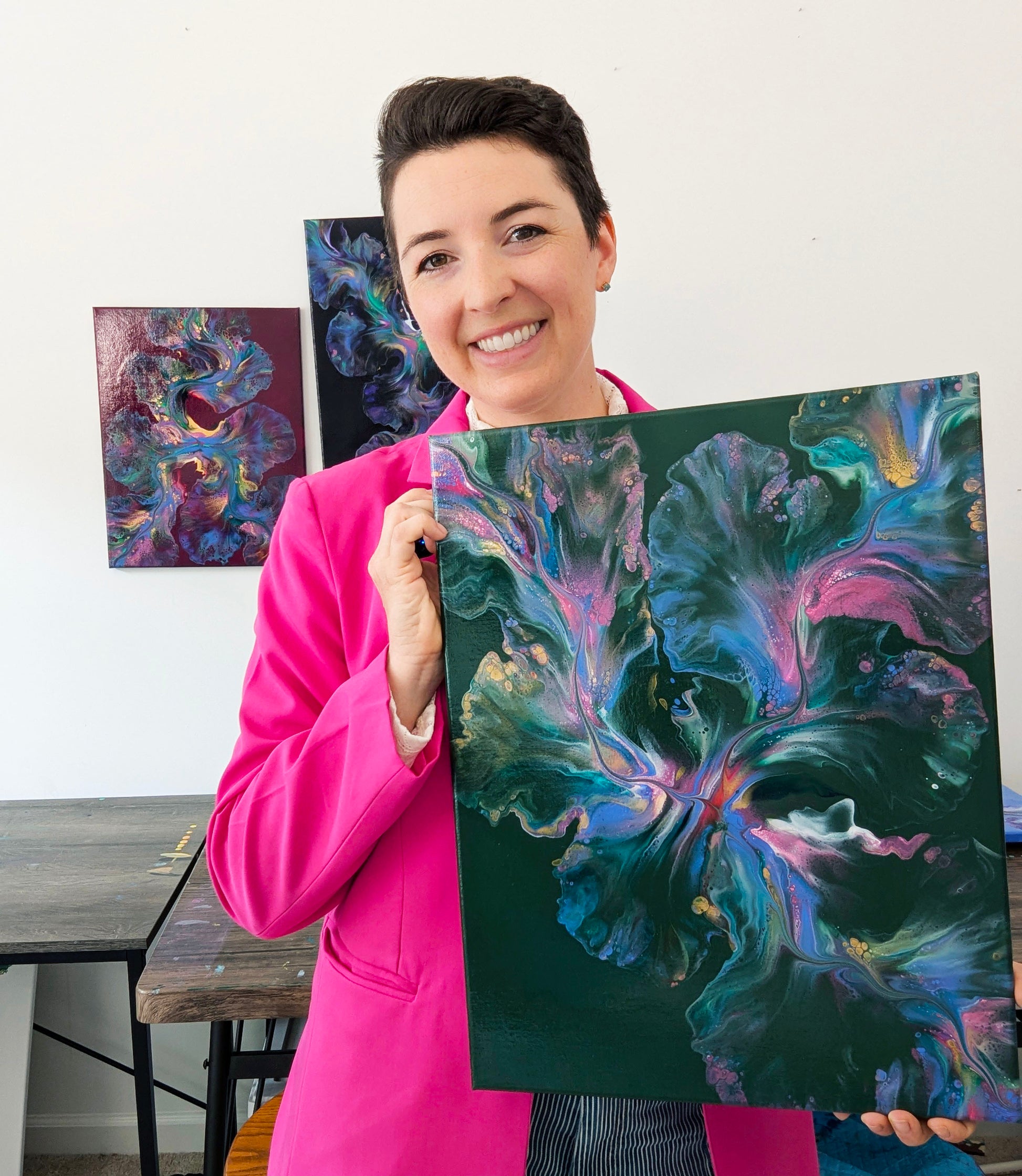 Artist Abby May in her studio in a bright pink blazer with green acrylic fluid art painting Enchanted Sky