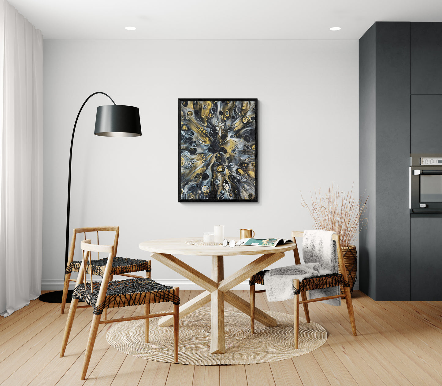 Abstract fluid art painting in black, gold, and gray tones framed in black, displayed in a modern dining area with a round wooden table, woven chairs, and a black floor lamp