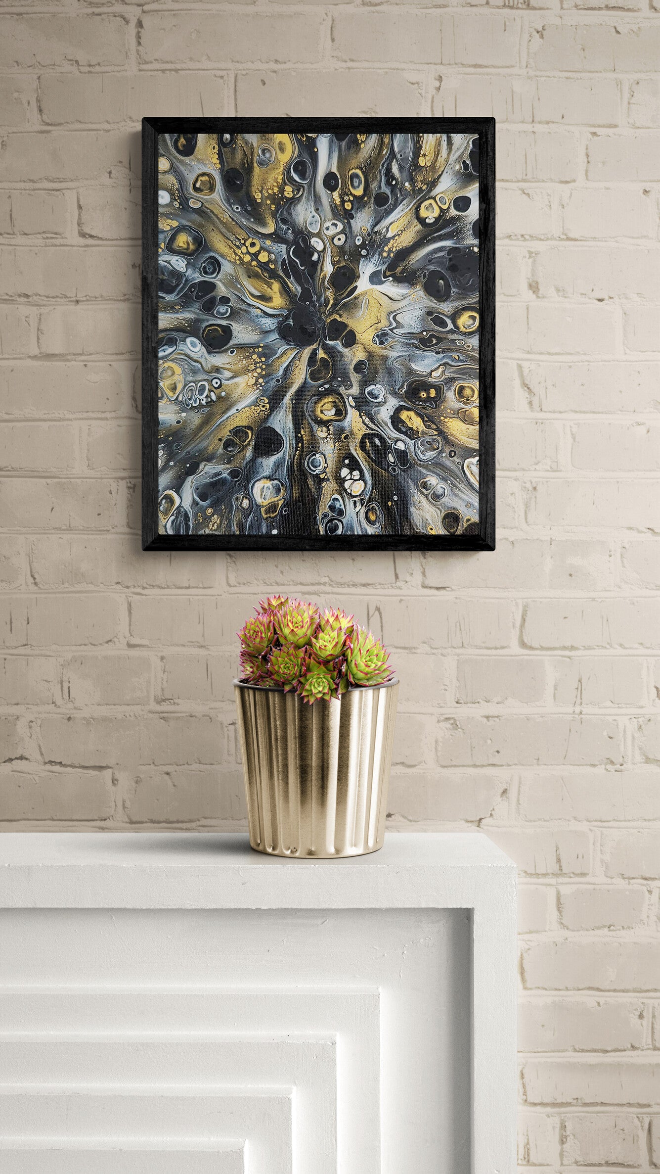 Close-up of a black-framed abstract fluid art print in gold, gray, and black tones displayed above a white mantel with a gold vase of succulents