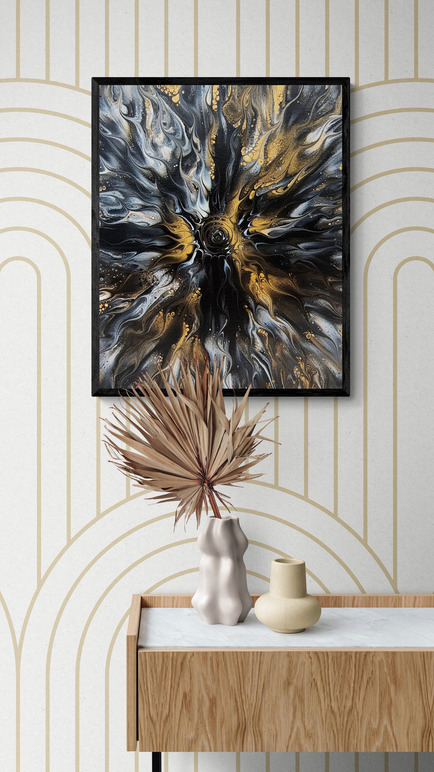 Abstract fluid art print in black, gold, and gray tones framed in black, hanging above a wood and marble console with sculptural vases and dried palm decor