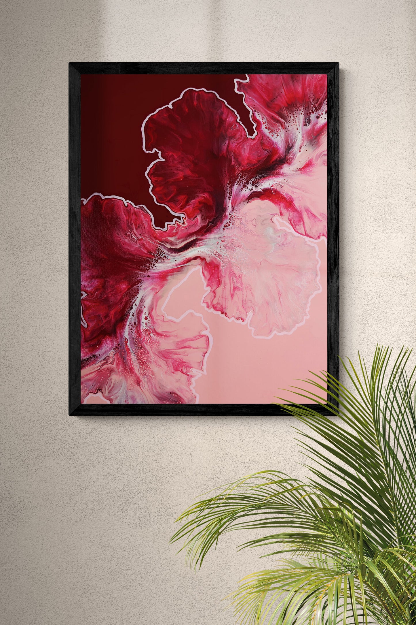 Close-up of a red and pink abstract fluid art print framed in black, displayed on a neutral wall with a green palm plant in the corner.