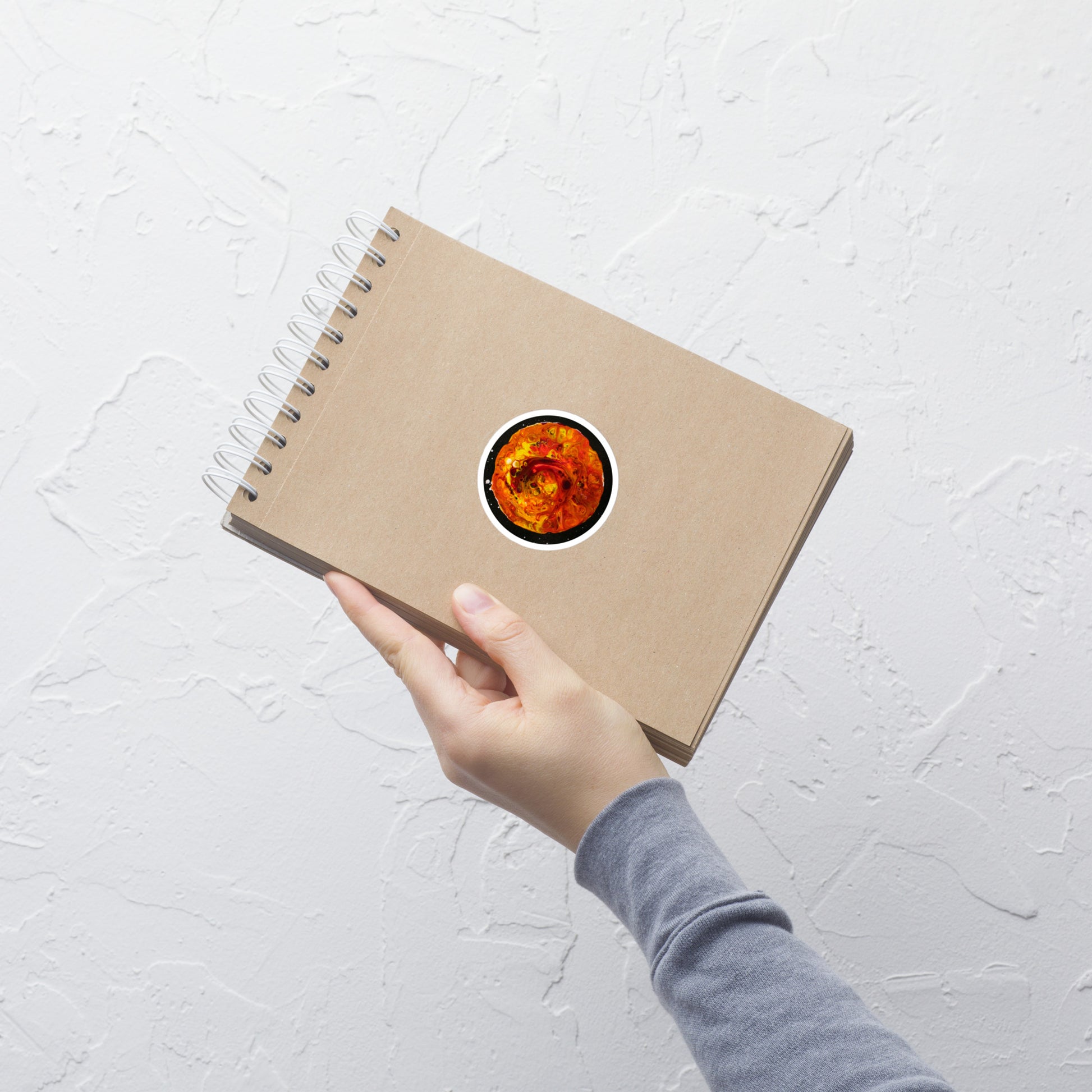 A hand holding a kraft spiral-bound notebook with an orange and red abstract moon sticker centered on the cover, shown against a white textured wall.