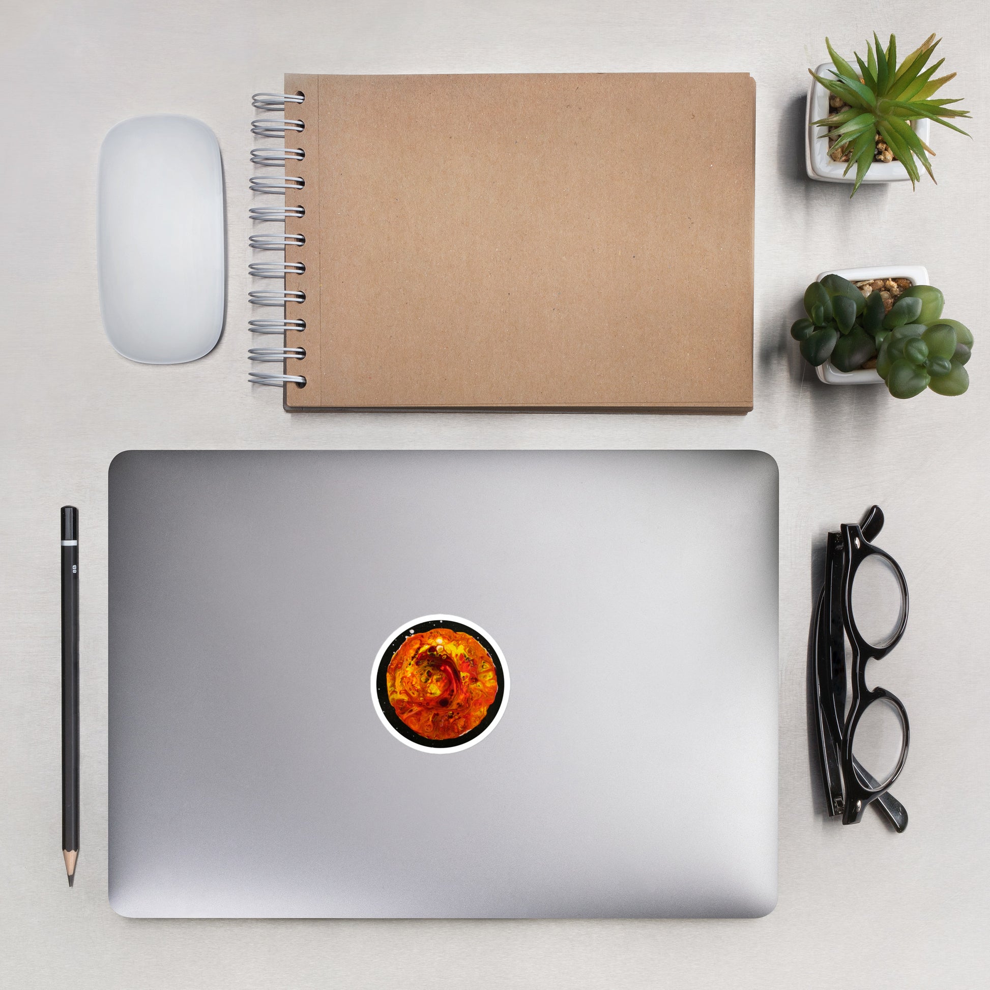 A flat lay of a workspace featuring a silver laptop with an orange and red moon sticker, a kraft notebook, black glasses, pencil, wireless mouse, and small potted succulents on a light gray surface.