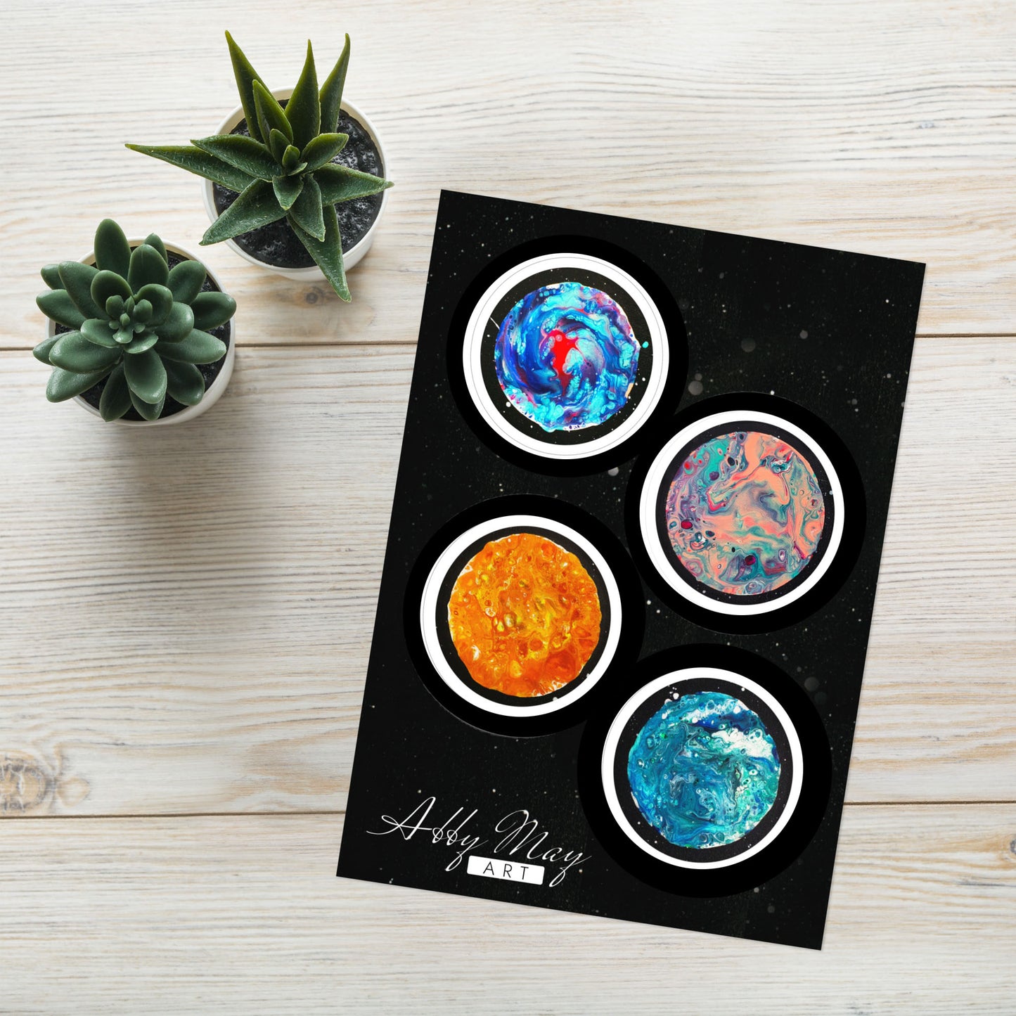 Sticker sheet displayed on a light wood surface next to two small succulent plants. The sheet includes four glossy circular stickers of colorful moons painted in a fluid abstract style, each with a black outline and white border.