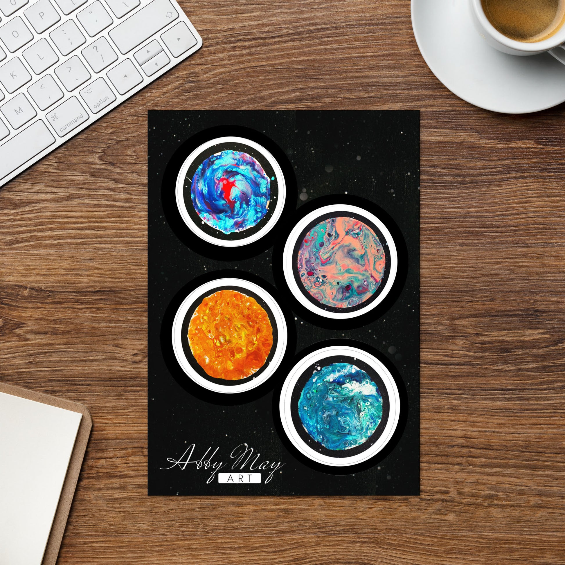 Sticker sheet placed on a dark wooden desk beside a white keyboard and a cup of coffee. Features four circular moon-themed abstract fluid art stickers with black outlines, designed by Abby May Art.