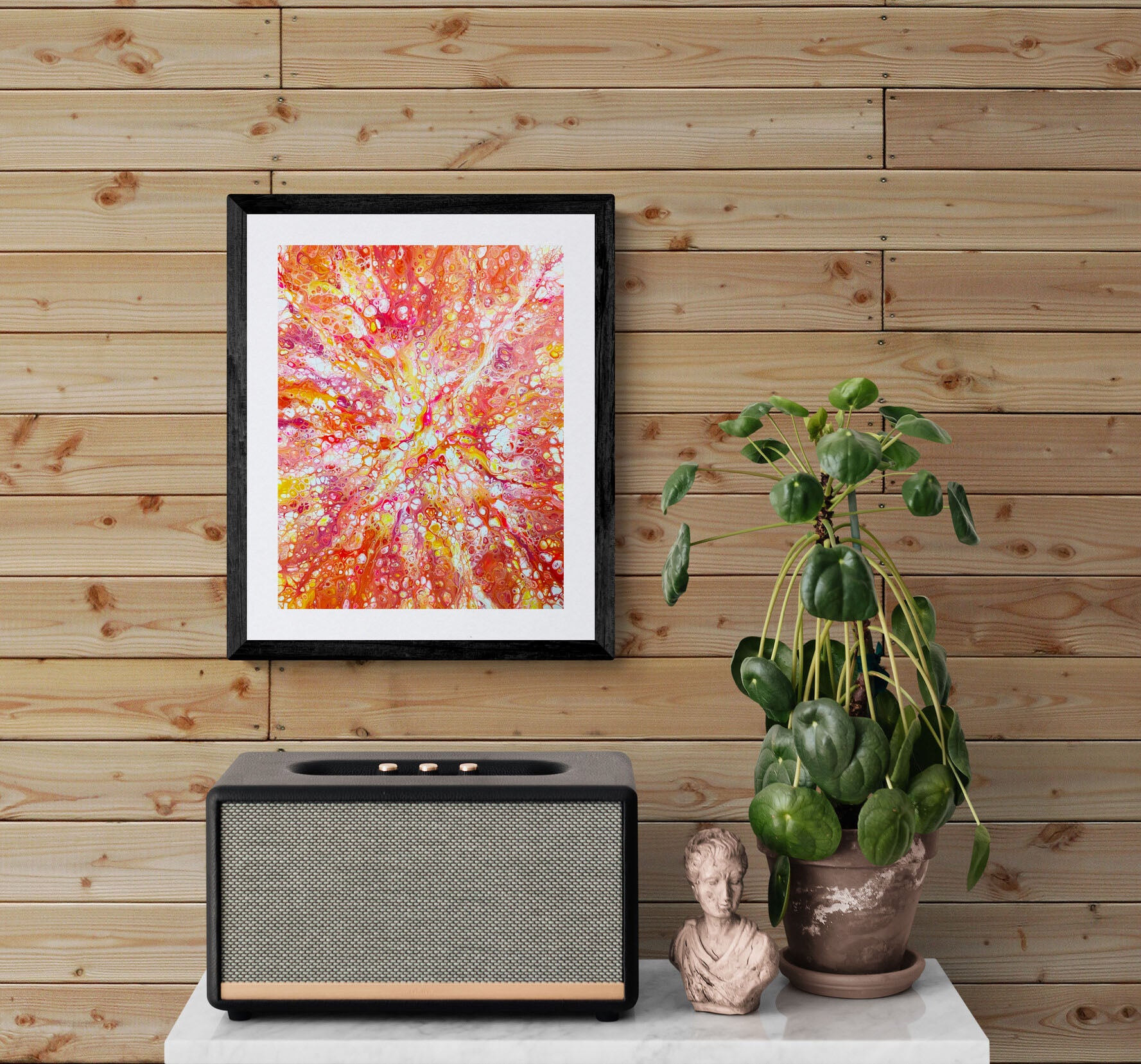 Vibrant “Phoenix Aflame” fluid art print in bold reds, oranges, and pinks, framed and hung on a wooden wall above a plant and modern speaker.
