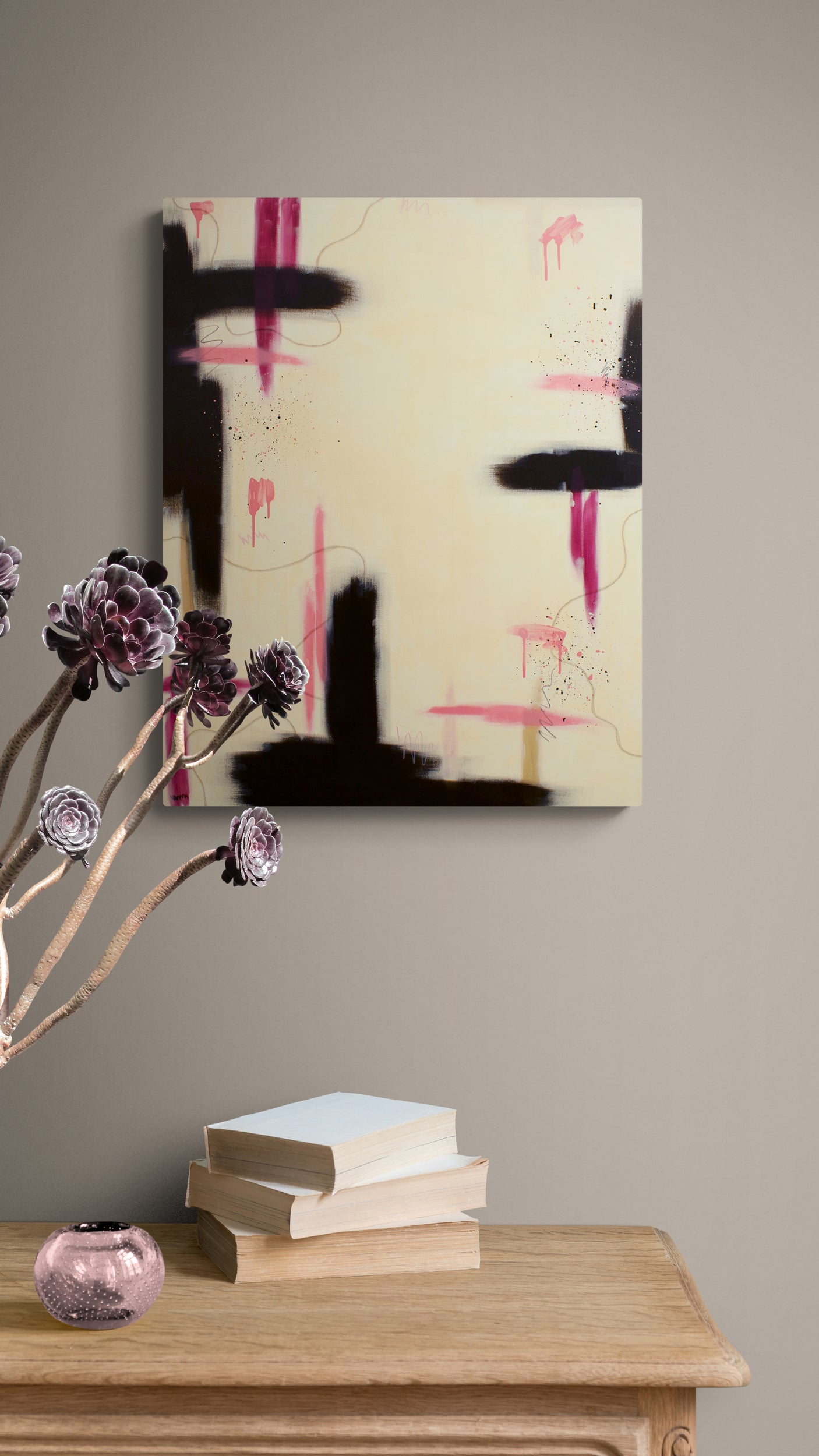 A large vertical contemporary abstract canvas print featuring bold black and vibrant pink brush strokes on a cream background, displayed on a taupe wall above a wooden console table with books and a decorative floral arrangement.