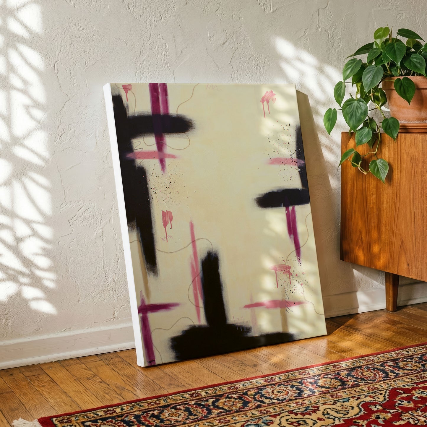 A large canvas print of the painting sitting on a wooden floor leaning against a textured white wall. There's a red patterned rug in front, and a wooden side table to the right with a terracotta potted plant.