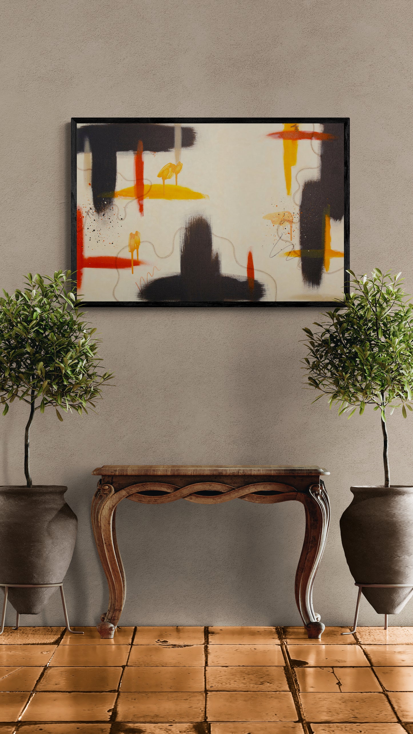 A framed horizontal abstract painting with raw umber, yellow, and red accents displayed in a black floater frame. The artwork is centered on a textured beige wall above an ornate wooden console table flanked by two large potted olive trees on a terracotta tiled floor.